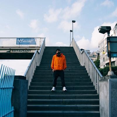 Dany Neville en veste orange en haut d'un escalier, devant une passerelle et des immeubles