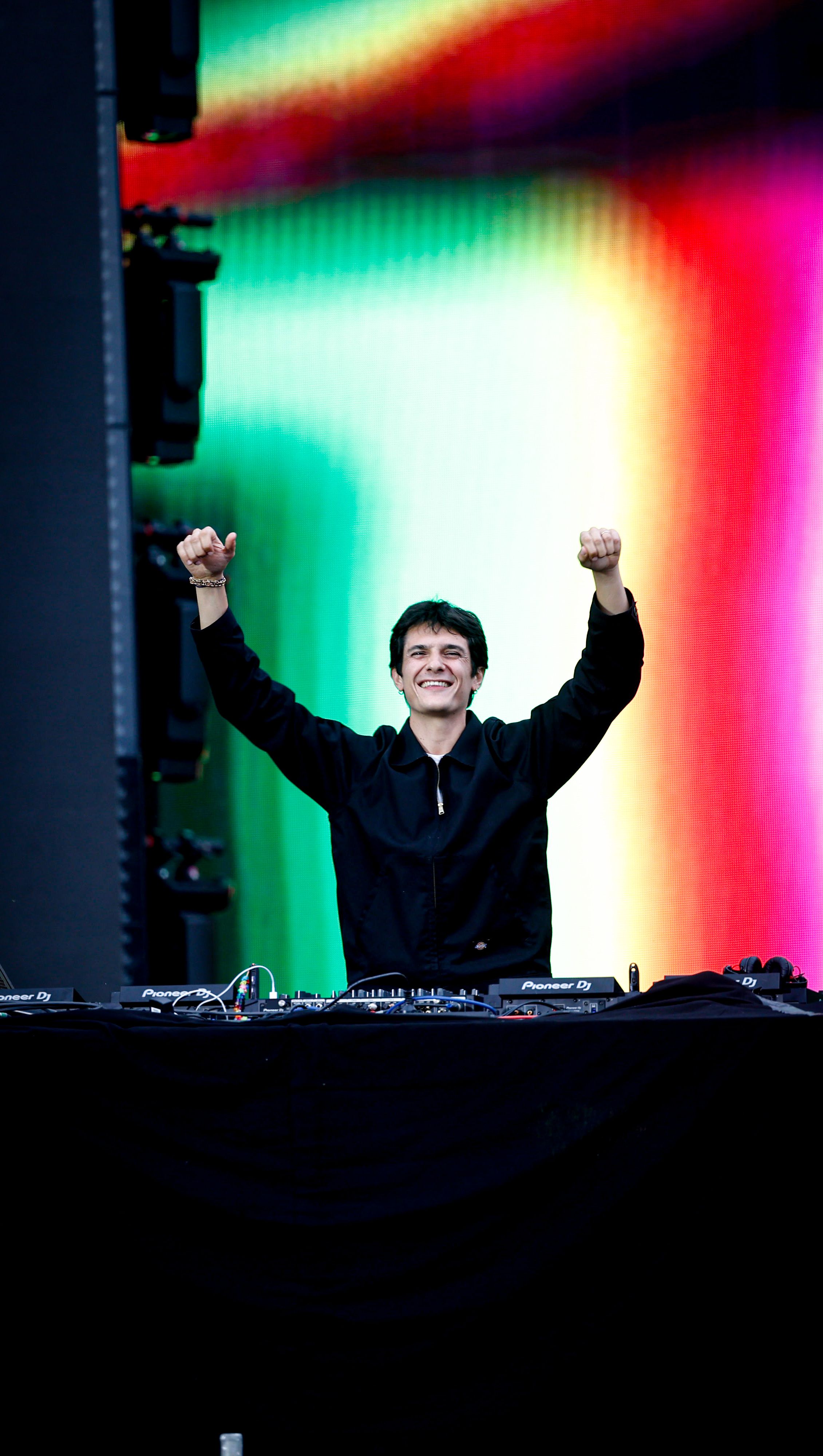 DJ souriant aux bras levés derrière ses platines, devant un écran coloré