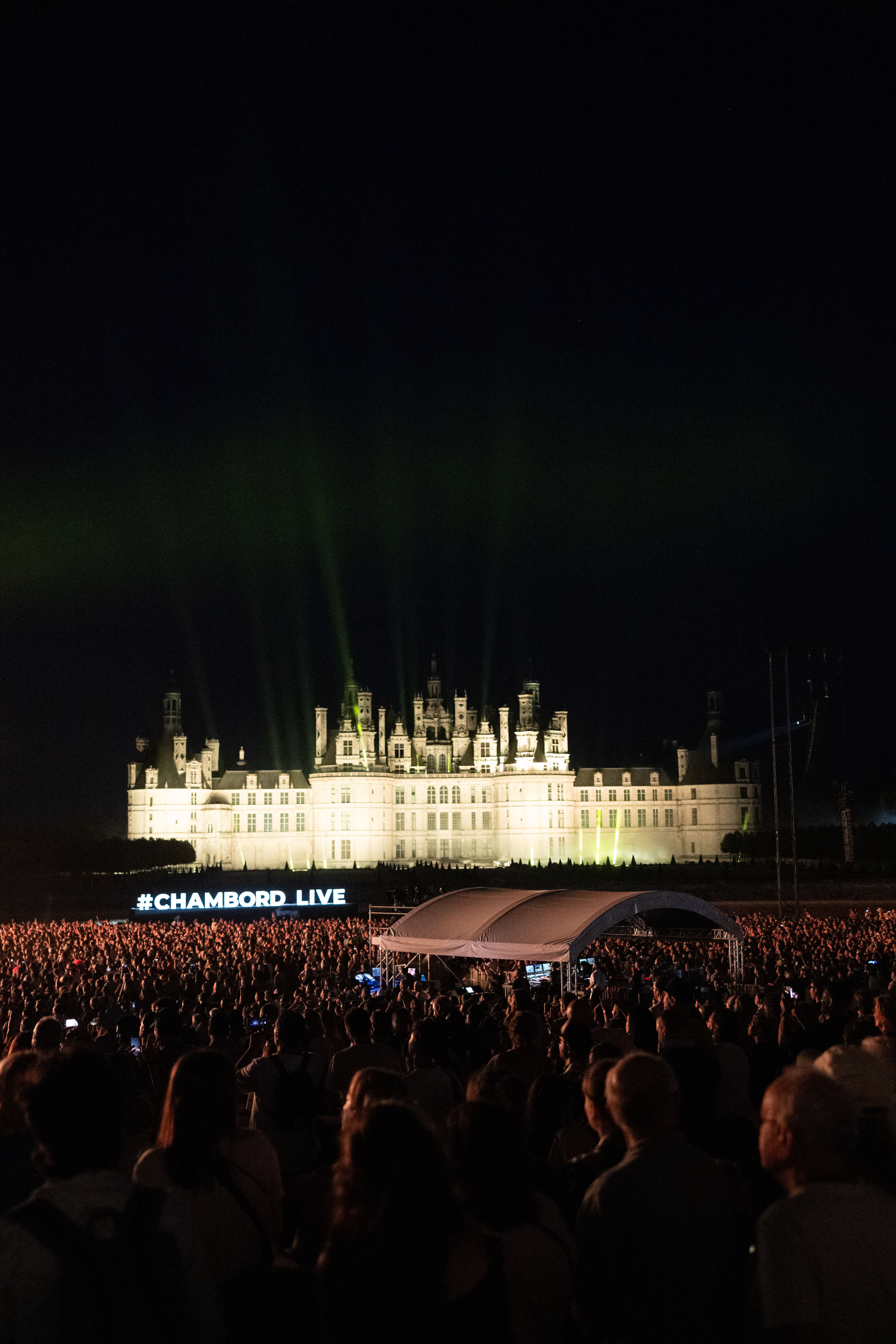 Foule dense face au château de Chambord illuminé de nuit, avec l'inscription #CHAMBORD LIVE au premier plan