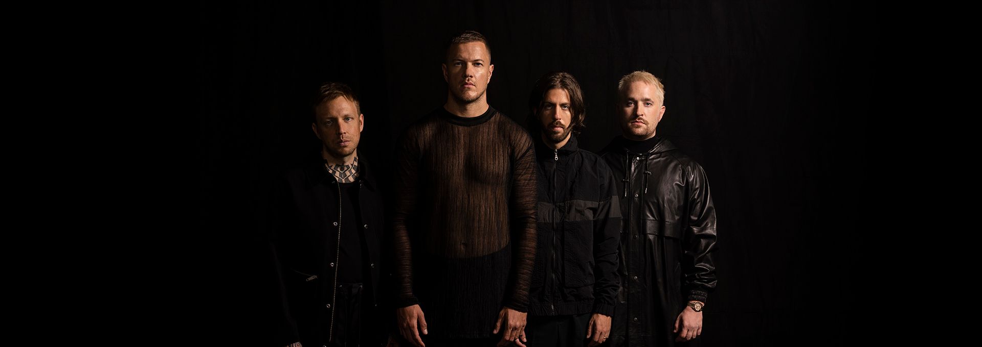 Portrait des quatre membres d'Imagine Dragons, vêtus de noir, sur fond sombre