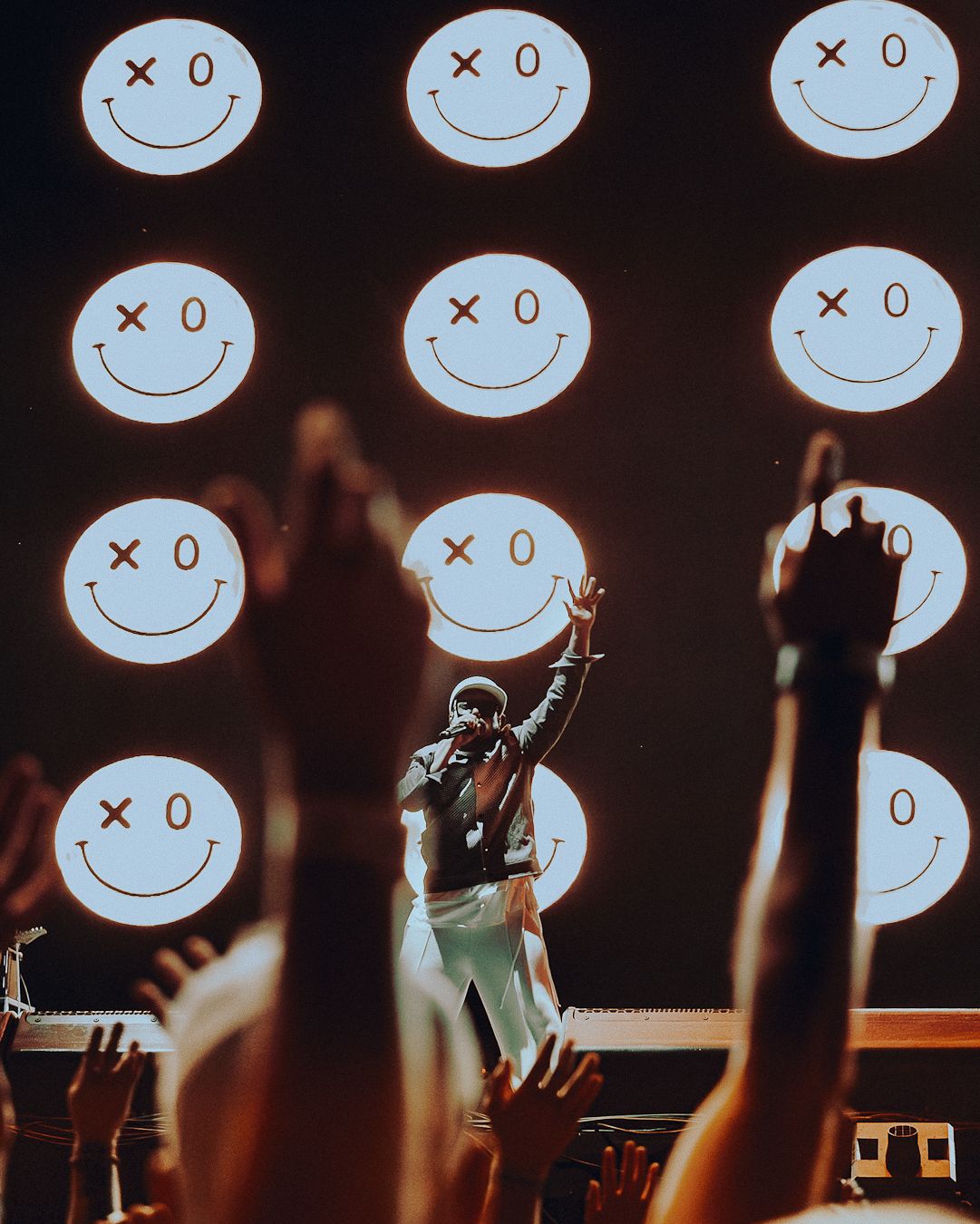 Artiste bras levé sur scène devant un écran de smileys, mains du public au premier plan