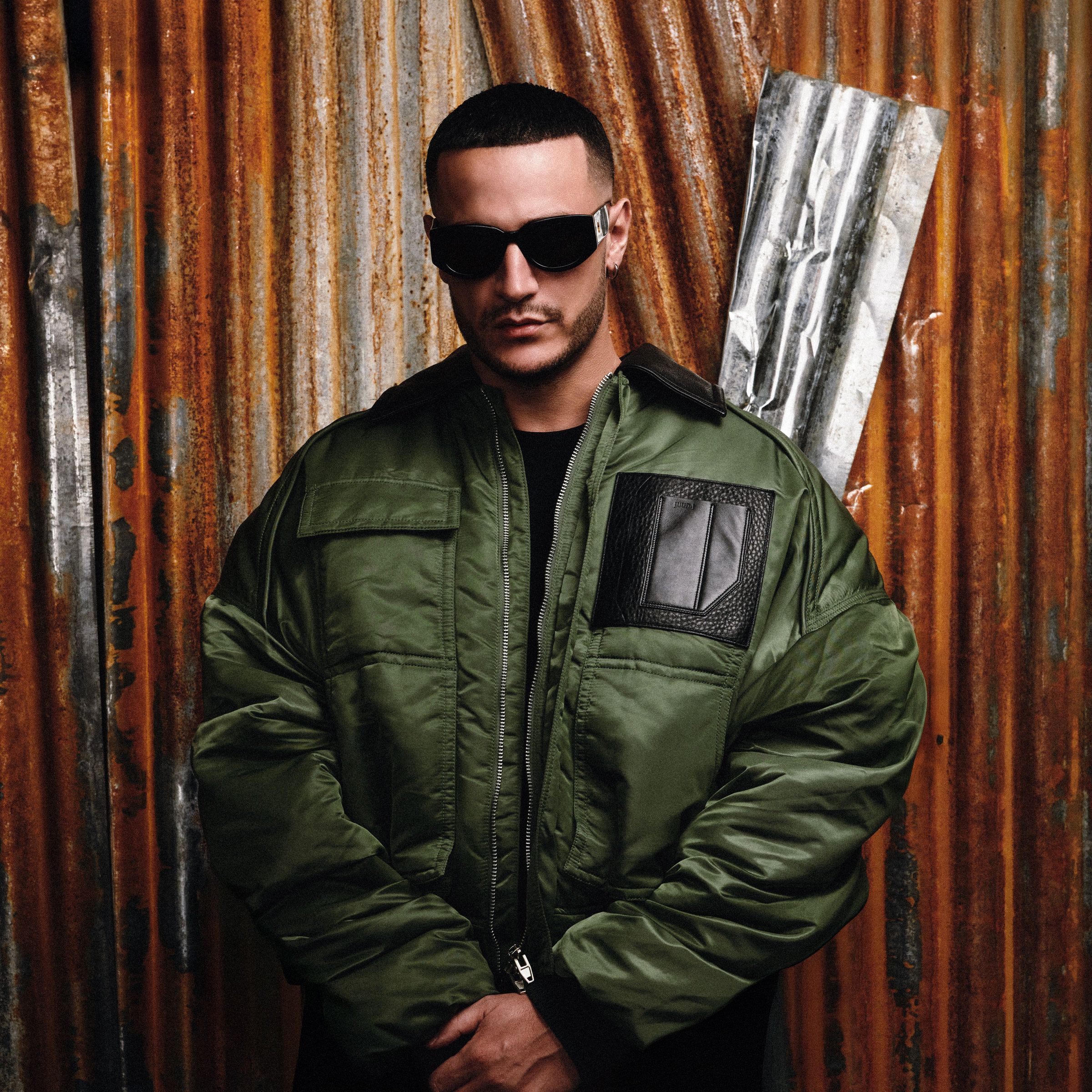 DJ Snake en blouson kaki et lunettes noires devant un mur de tôle rouillée