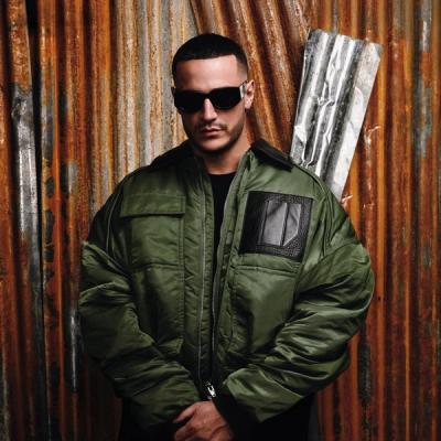 DJ Snake en blouson kaki et lunettes noires devant un mur de tôle rouillée