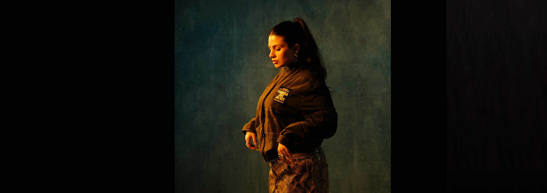 Eve La Marka de profil, queue de cheval et veste sombre, sur fond bleu-vert éclairé de lumière chaude