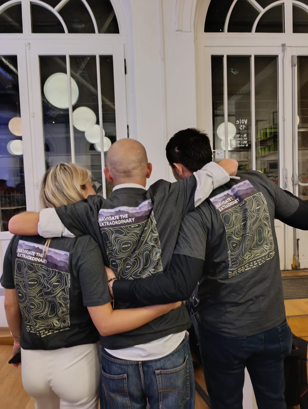 Trois membres de l'équipe TOOGO de dos, bras dessus bras dessous, portant le T-shirt Navigate the Extraordinary.