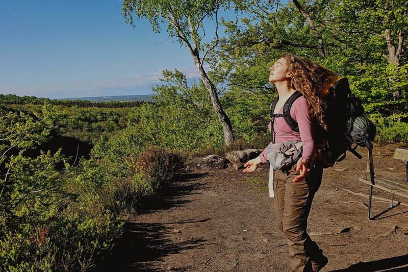 A hiker with a backpack pauses on a sunlit trail at a forest overlook in Söderåsen National Park, Sweden, face turned up to the sun above a wide green ravine landscape