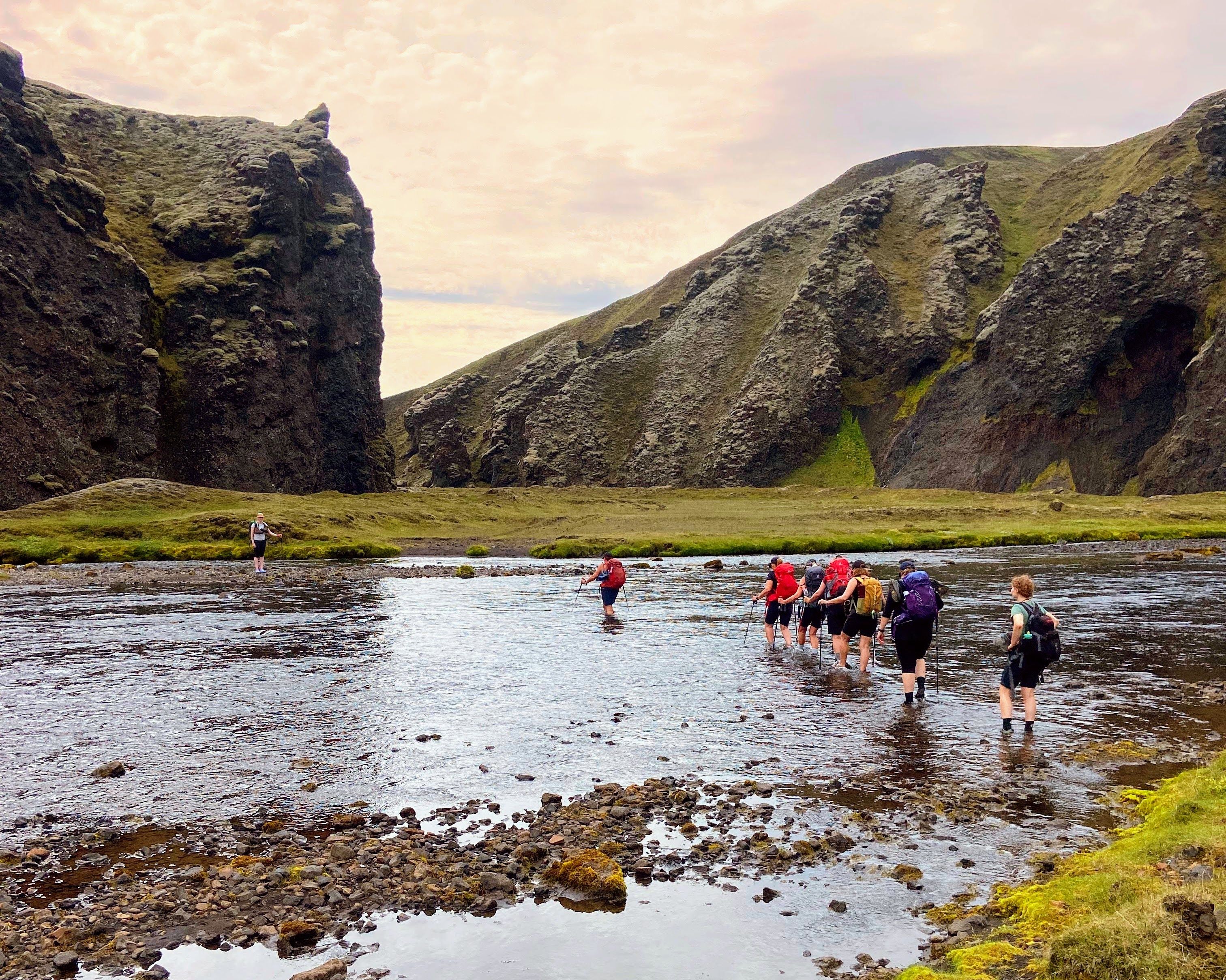 A line of hikers crosses a wide gravel plain toward highland mountains under blue sky