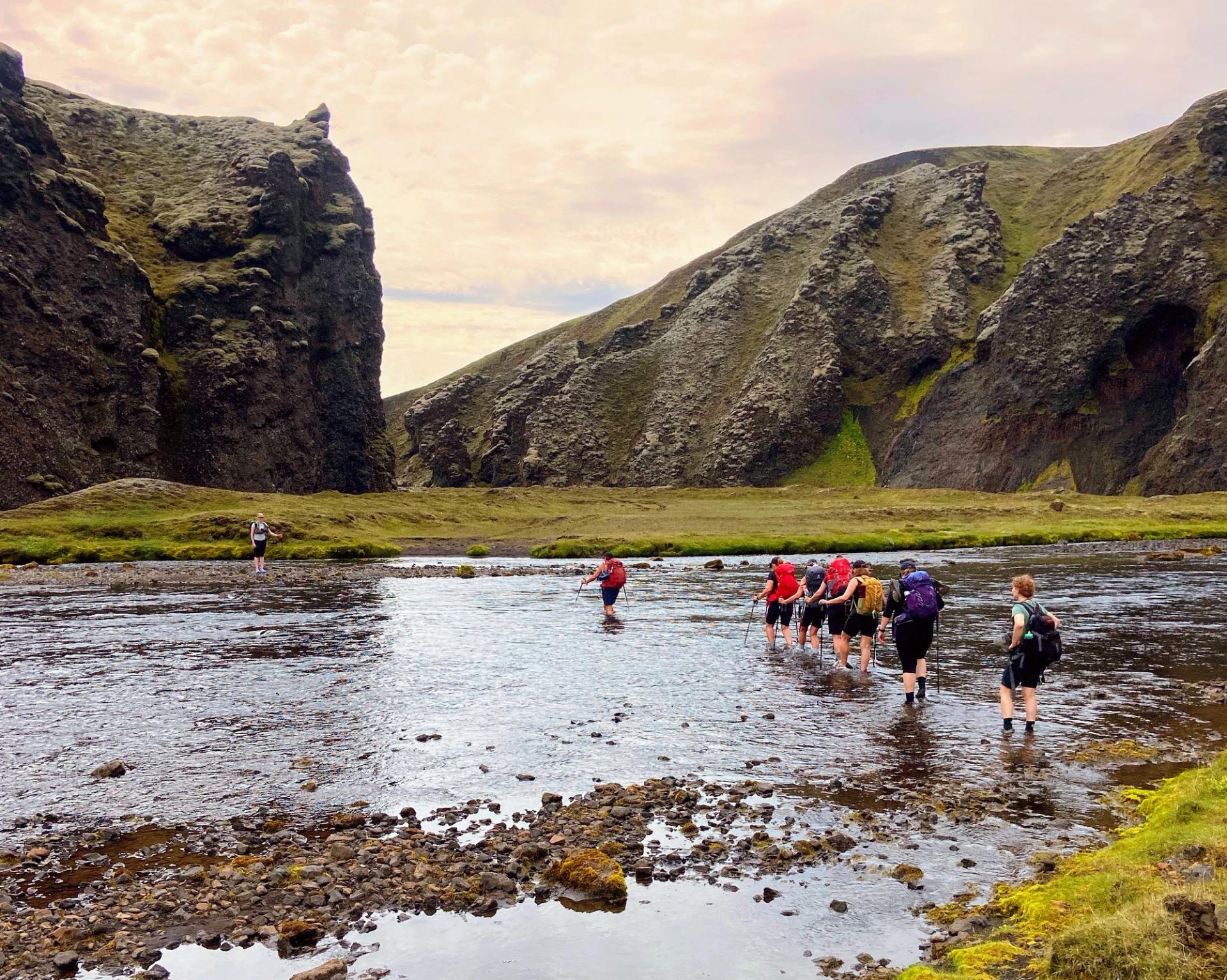 A line of hikers crosses a wide gravel plain toward highland mountains under blue sky