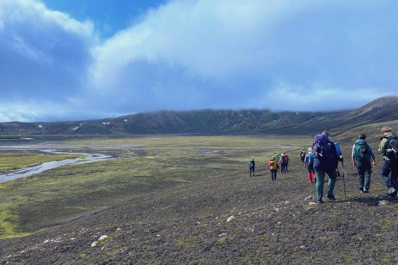 A line of hikers crosses a wide gravel plain toward highland mountains under blue sky