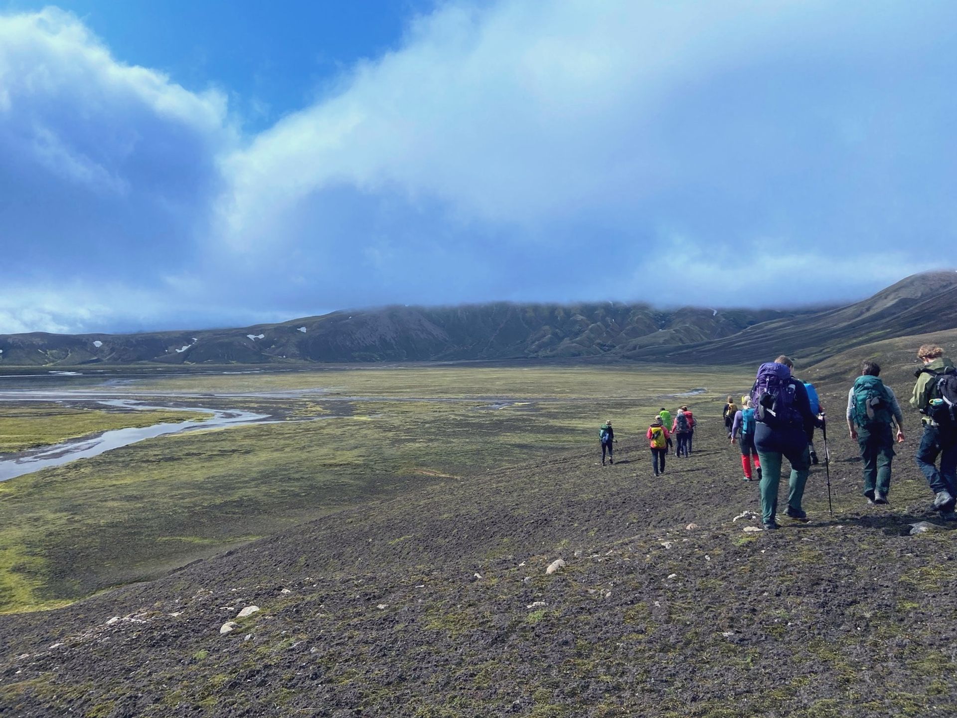 A line of hikers crosses a wide gravel plain toward highland mountains under blue sky