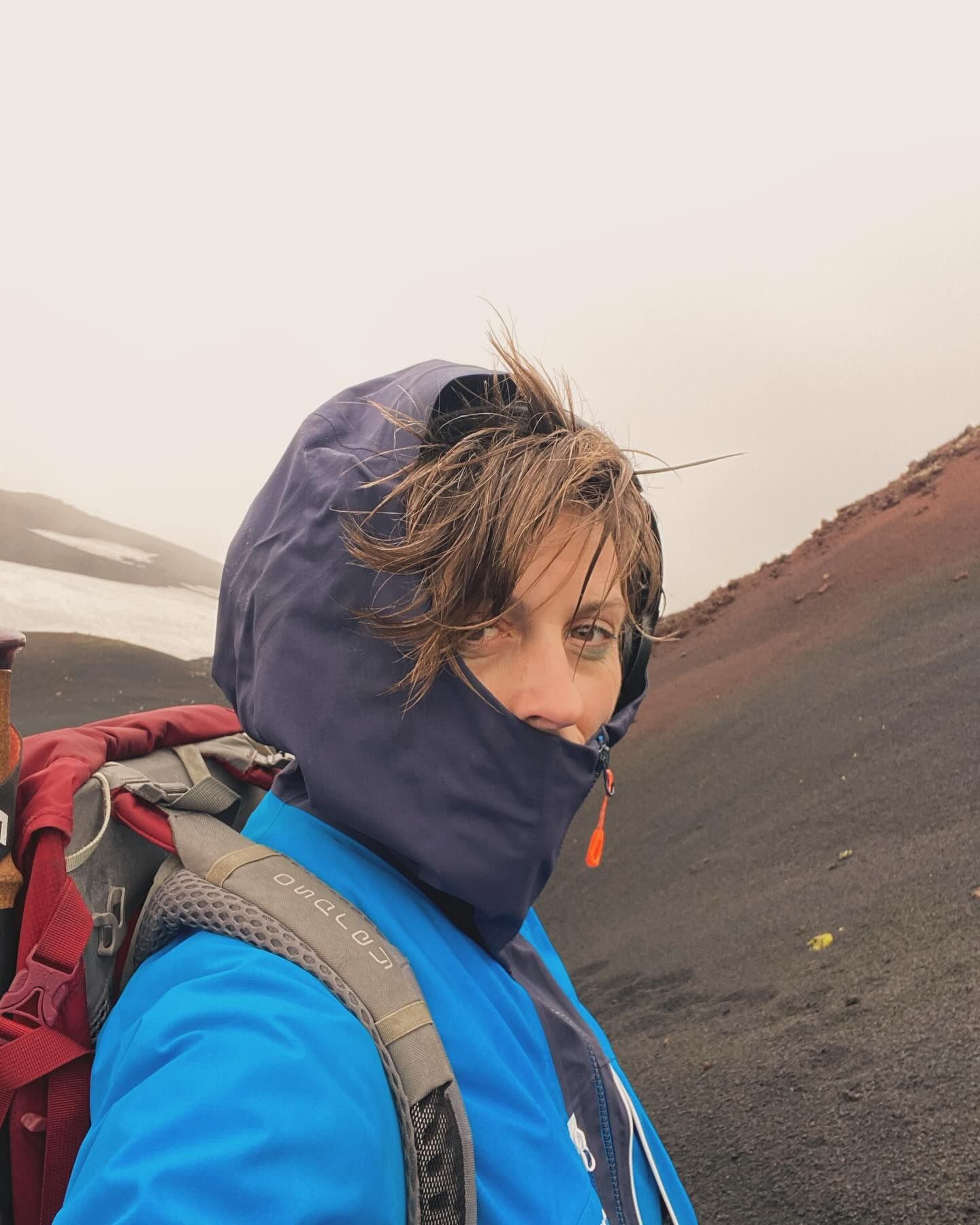A hiker in a blue jacket and hood, hair windblown, on a misty black volcanic slope with a patch of snow behind.