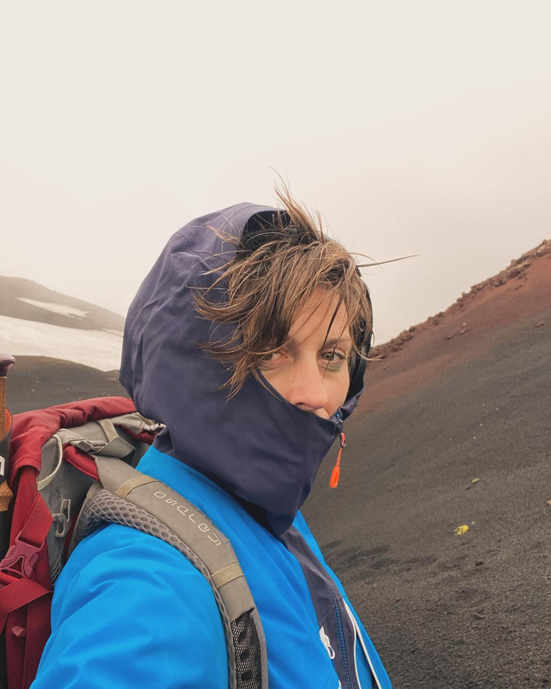A hiker in a blue jacket and hood, hair windblown, on a misty black volcanic slope with a patch of snow behind.