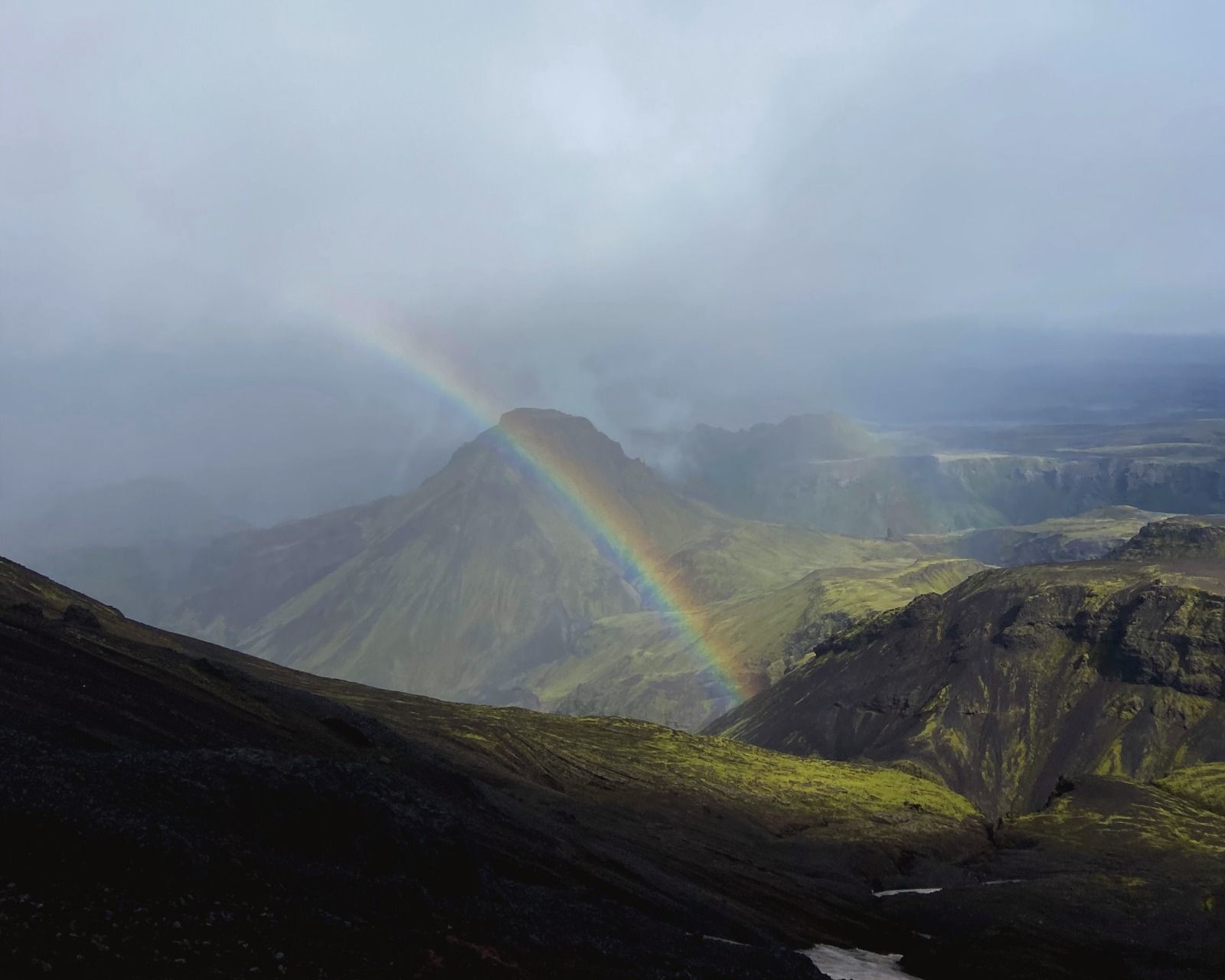 A vivid rainbow arches over dark green and rhyolite mountains in the Icelandic highlands