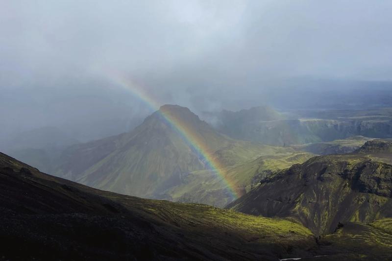 A vivid rainbow arches over dark green and rhyolite mountains in the Icelandic highlands