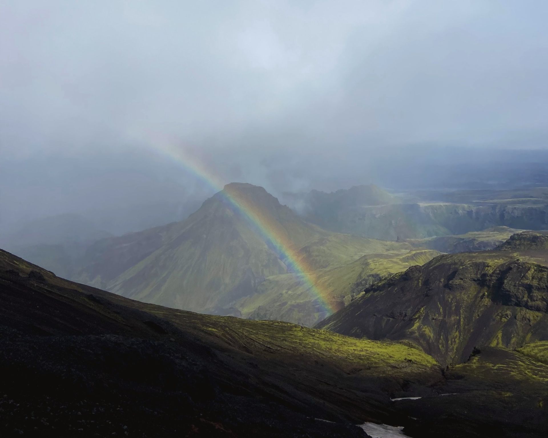 A vivid rainbow arches over dark green and rhyolite mountains in the Icelandic highlands