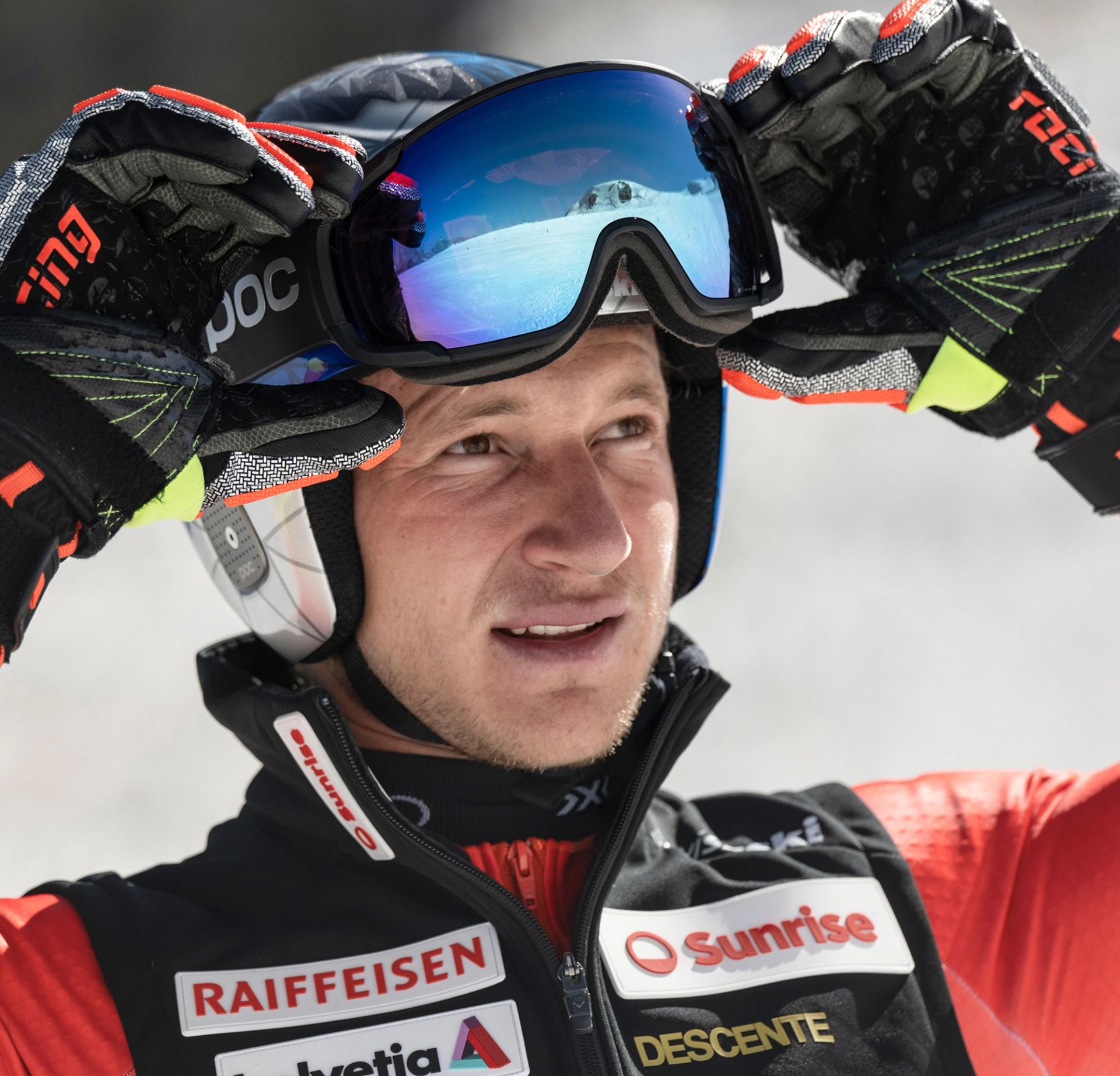 A skier wearing a helmet, reflective goggles, and padded gloves adjusts the sides of their goggles. The reflection in the lens shows a snowy mountain landscape. The skier is dressed in a black and red racing suit covered with sponsor logos