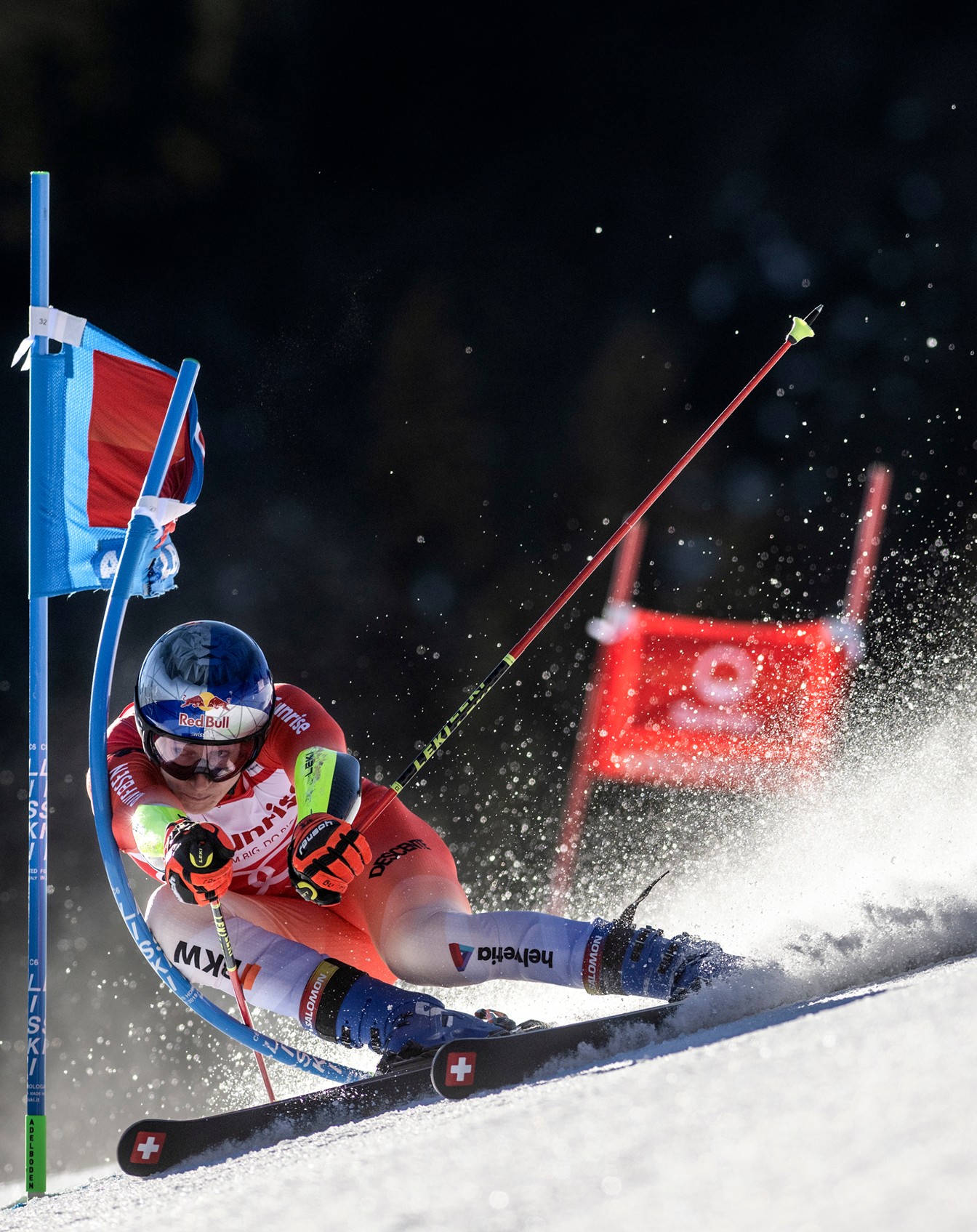 A skier in a red racing suit carves through slalom course on a groomed snowy slope. Orange gates line the path while blue dye marks the turns. 
