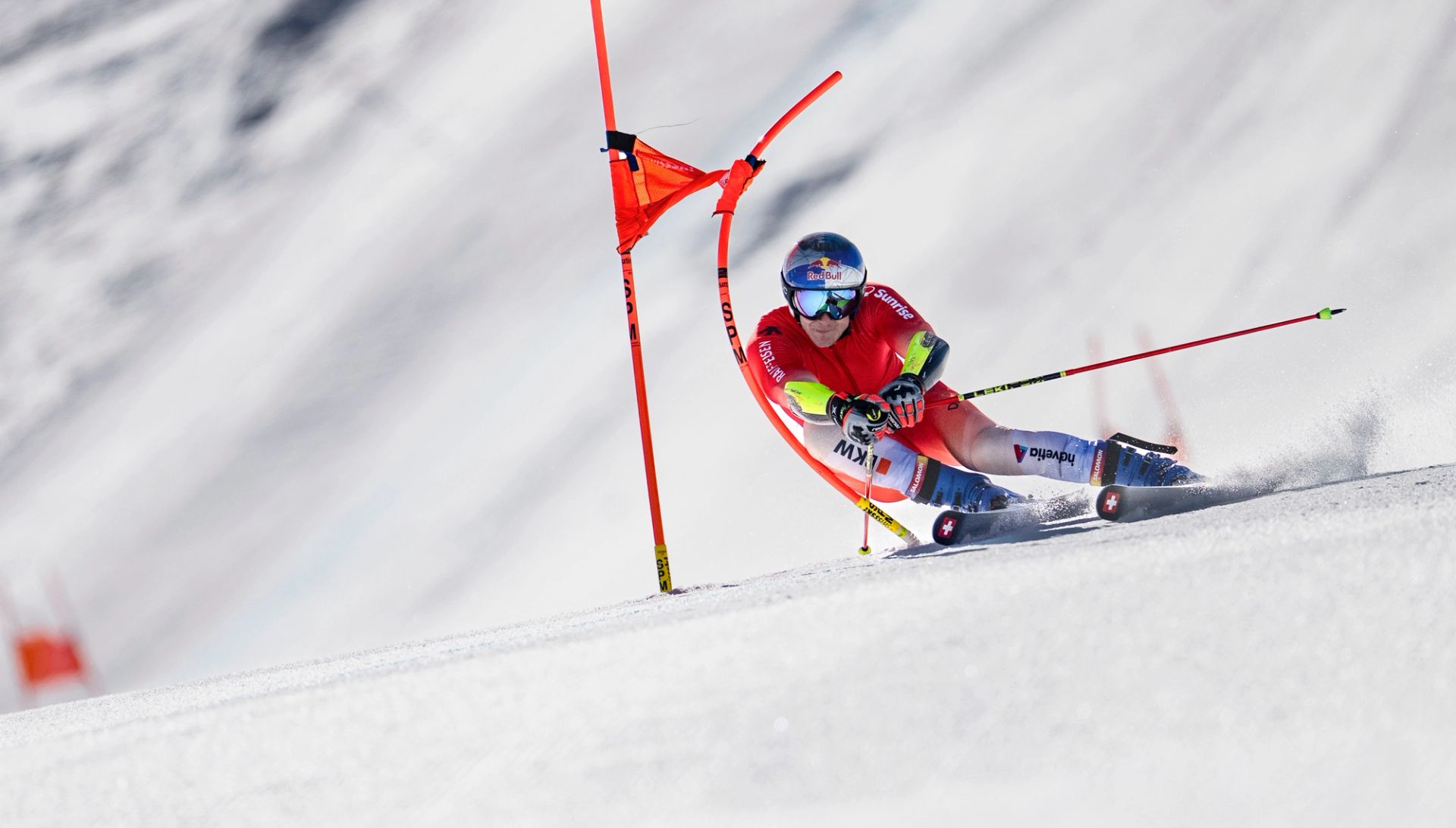 Marco Odermatt in a racing suit and helmet carving through a slalom course, leaning into a turn with poles and snow spray visible