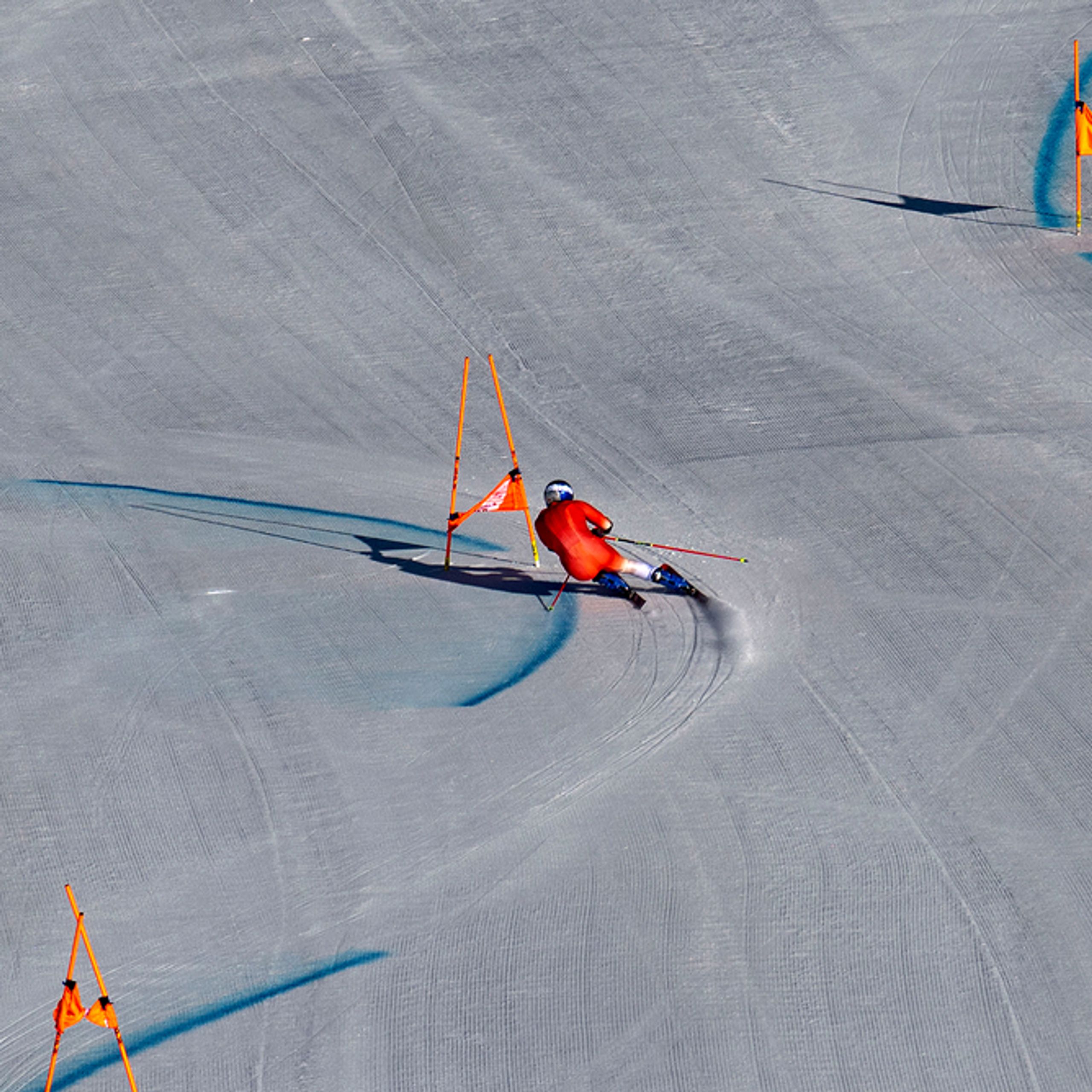 A skier in a red racing suit carves through a giant slalom course on a groomed snowy slope. Orange gates line the path while blue dye marks the turns. The shot is taken from far above, emphasizing the wide, open terrain