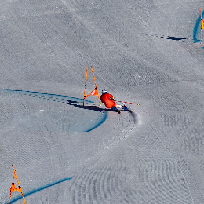 A skier in a red racing suit carves through a giant slalom course on a groomed snowy slope. Orange gates line the path while blue dye marks the turns. The shot is taken from far above, emphasizing the wide, open terrain