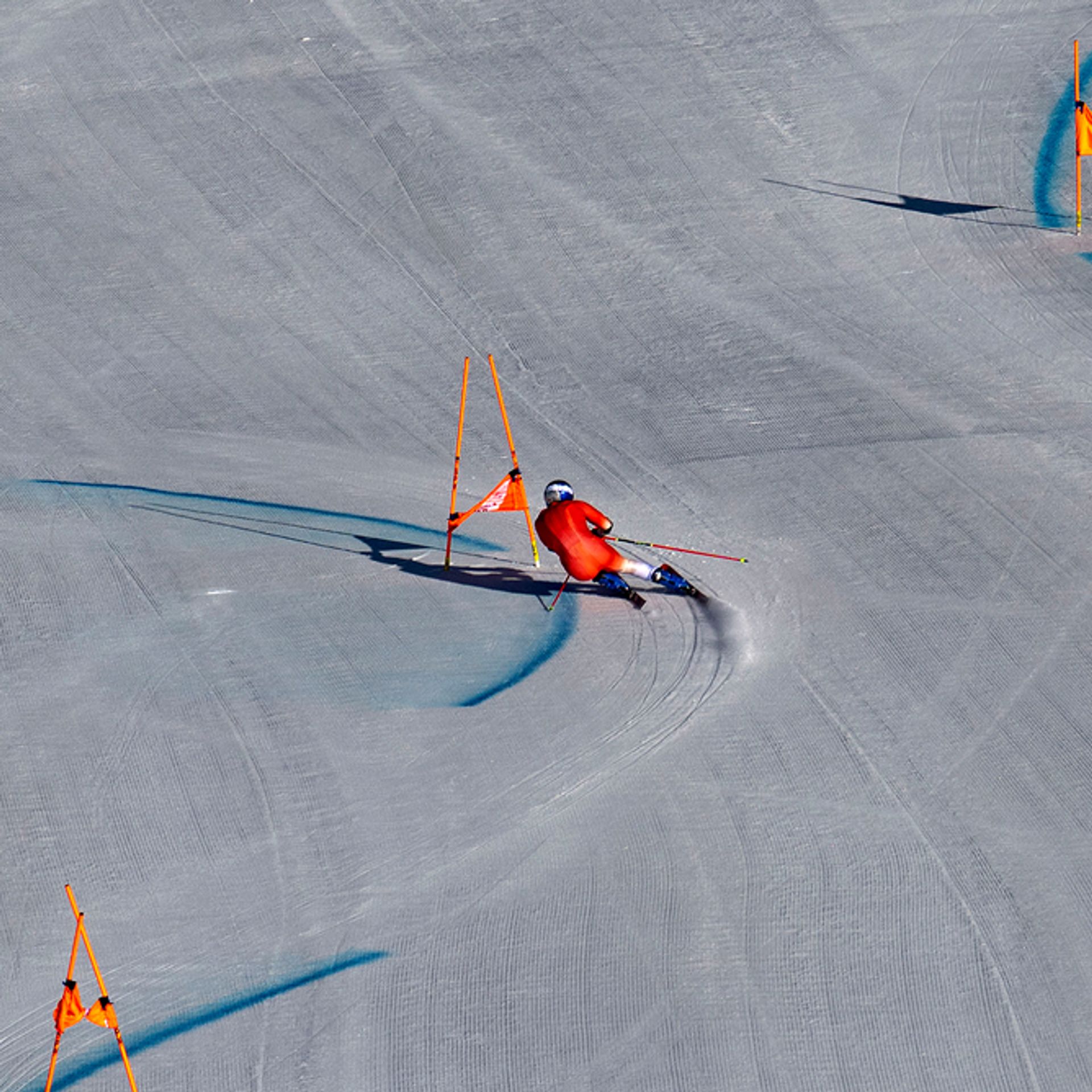 A skier in a red racing suit carves through a giant slalom course on a groomed snowy slope. Orange gates line the path while blue dye marks the turns. The shot is taken from far above, emphasizing the wide, open terrain