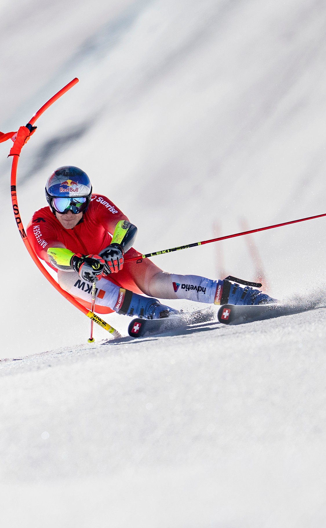 Marco Odermatt in a racing suit and helmet carving through a slalom course, leaning into a turn with poles and snow spray visible