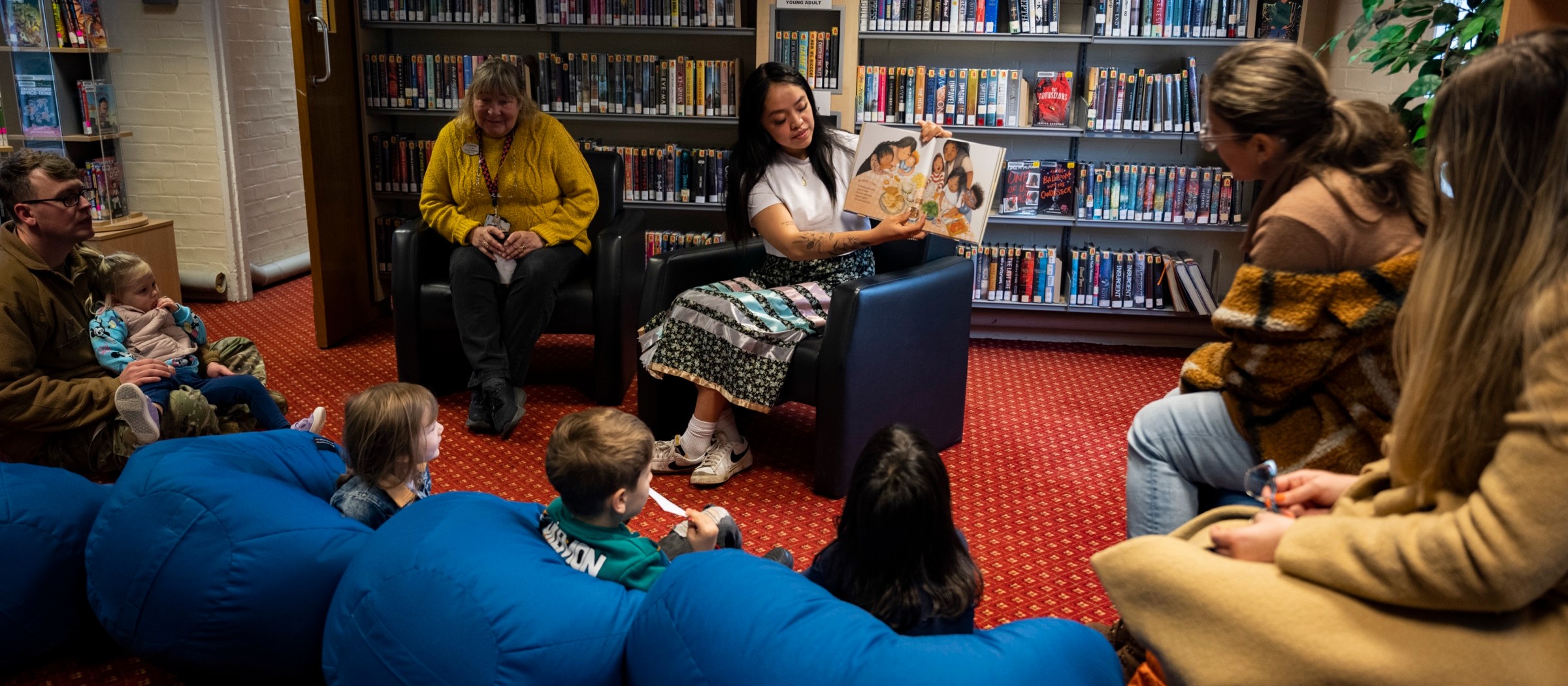 Una mujer lee un libro ilustrado a niños pequeños sentados en pufs en una biblioteca