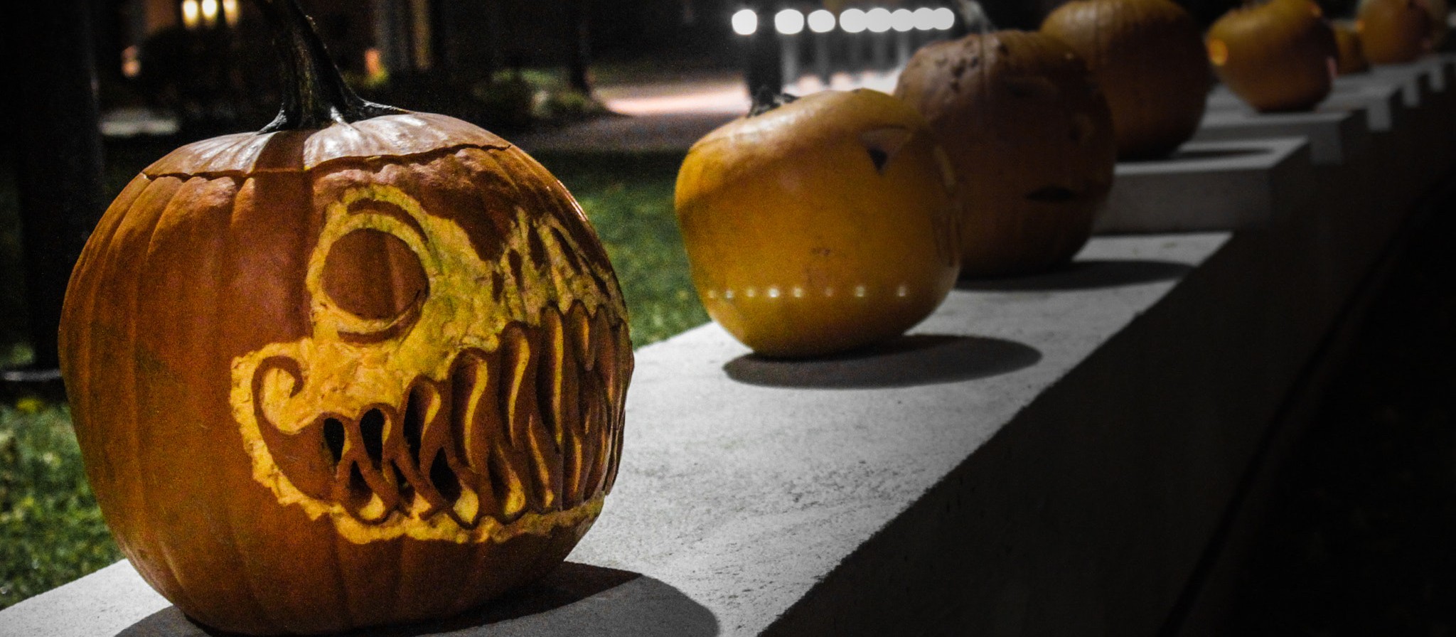 Una fila de calabazas talladas sobre un muro bajo de noche, con una calabaza de dientes afilados al frente