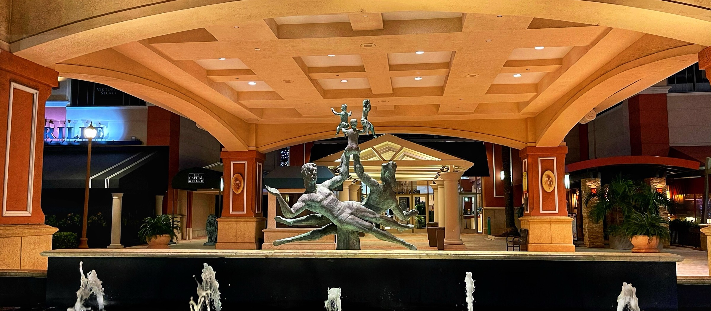 The arched entrance of Galleria Fort Lauderdale at night, with a bronze sculpture over a lit fountain