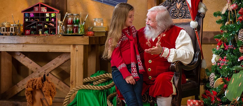 Una niña con suéter rojo sentada junto a Santa en su taller de madera