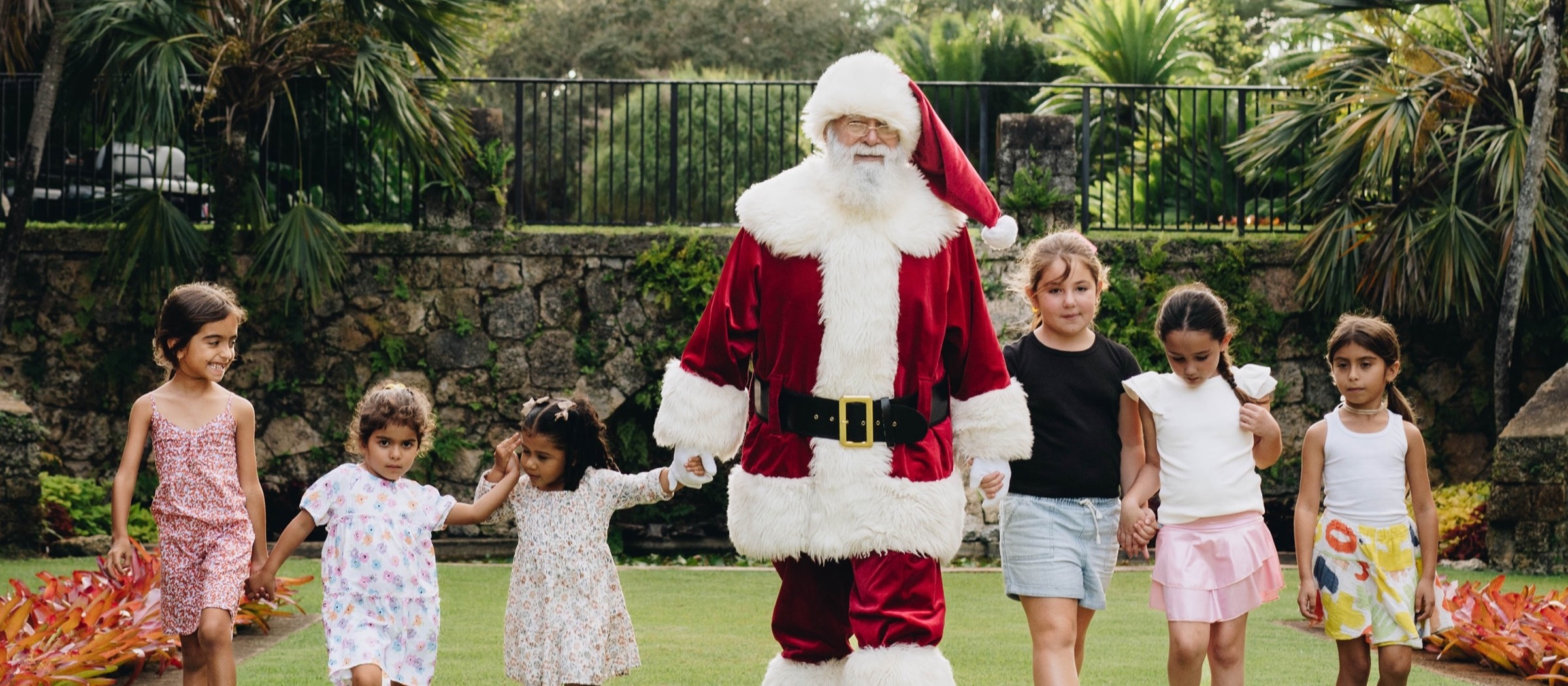 Santa Claus walking across a garden lawn holding hands with a group of children