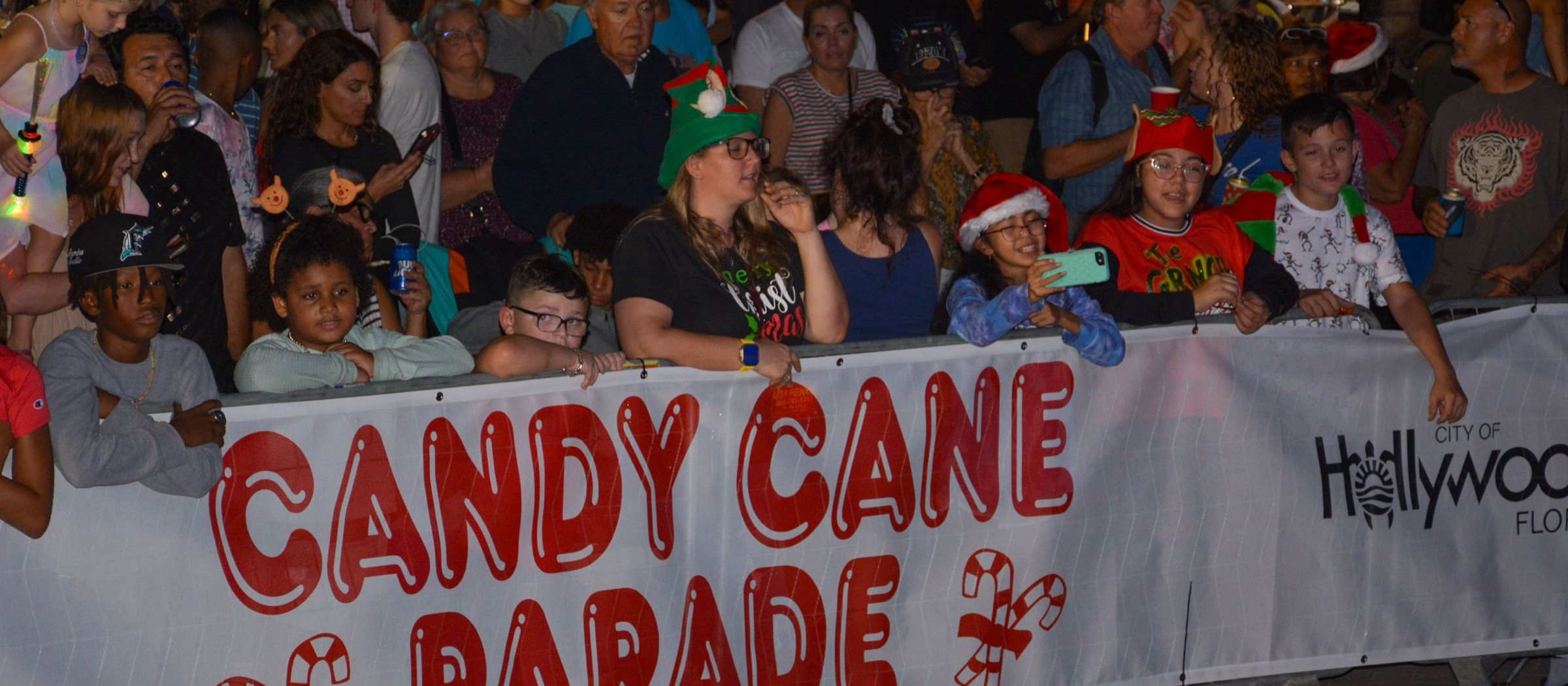 Niños con gorros de Santa y familias mirando el desfile detrás de una pancarta del Candy Cane Parade de noche