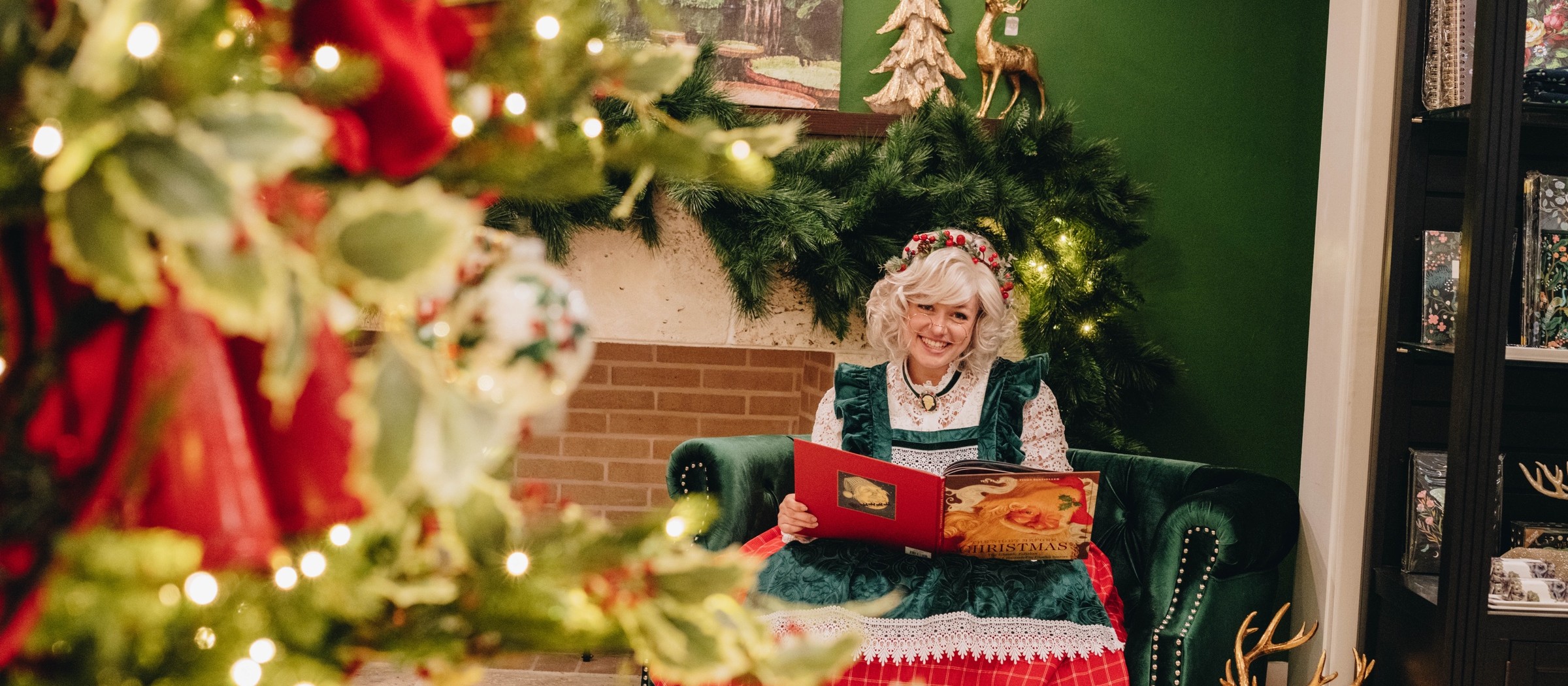 Mrs. Claus reading a storybook in an armchair by a fireplace, a decorated tree in the foreground
