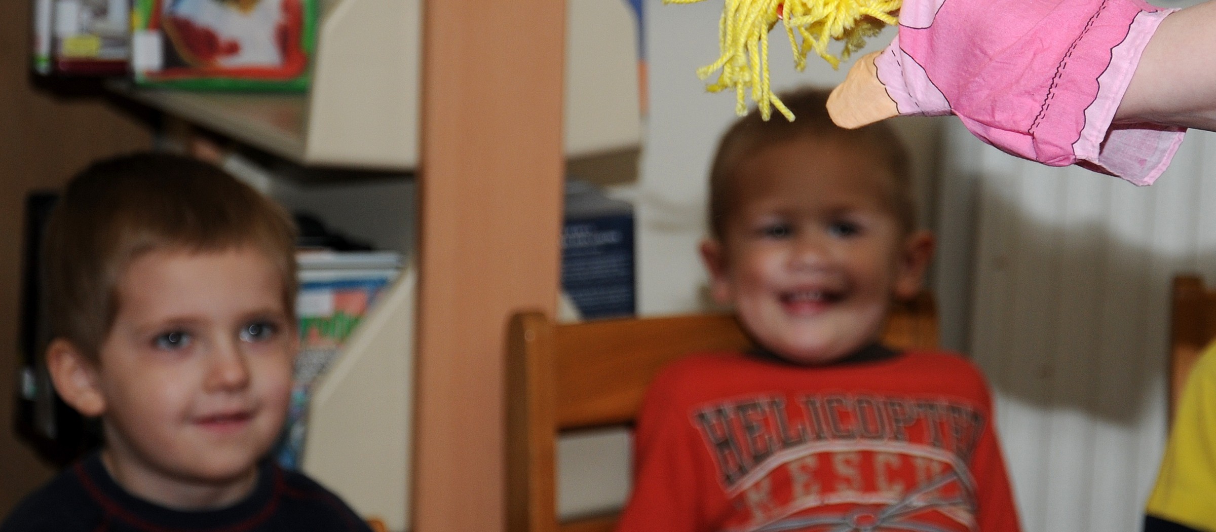 Two young boys at a library smile at a pink glove puppet with yellow yarn hair held up by a storyteller