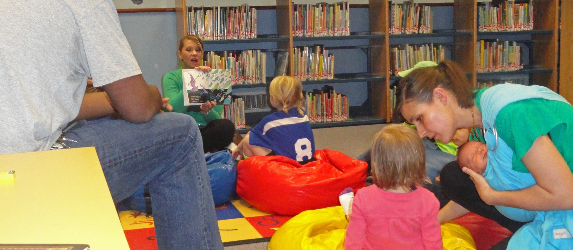 Niños pequeños en pufs de colores y una madre con su bebé durante una hora del cuento en la biblioteca