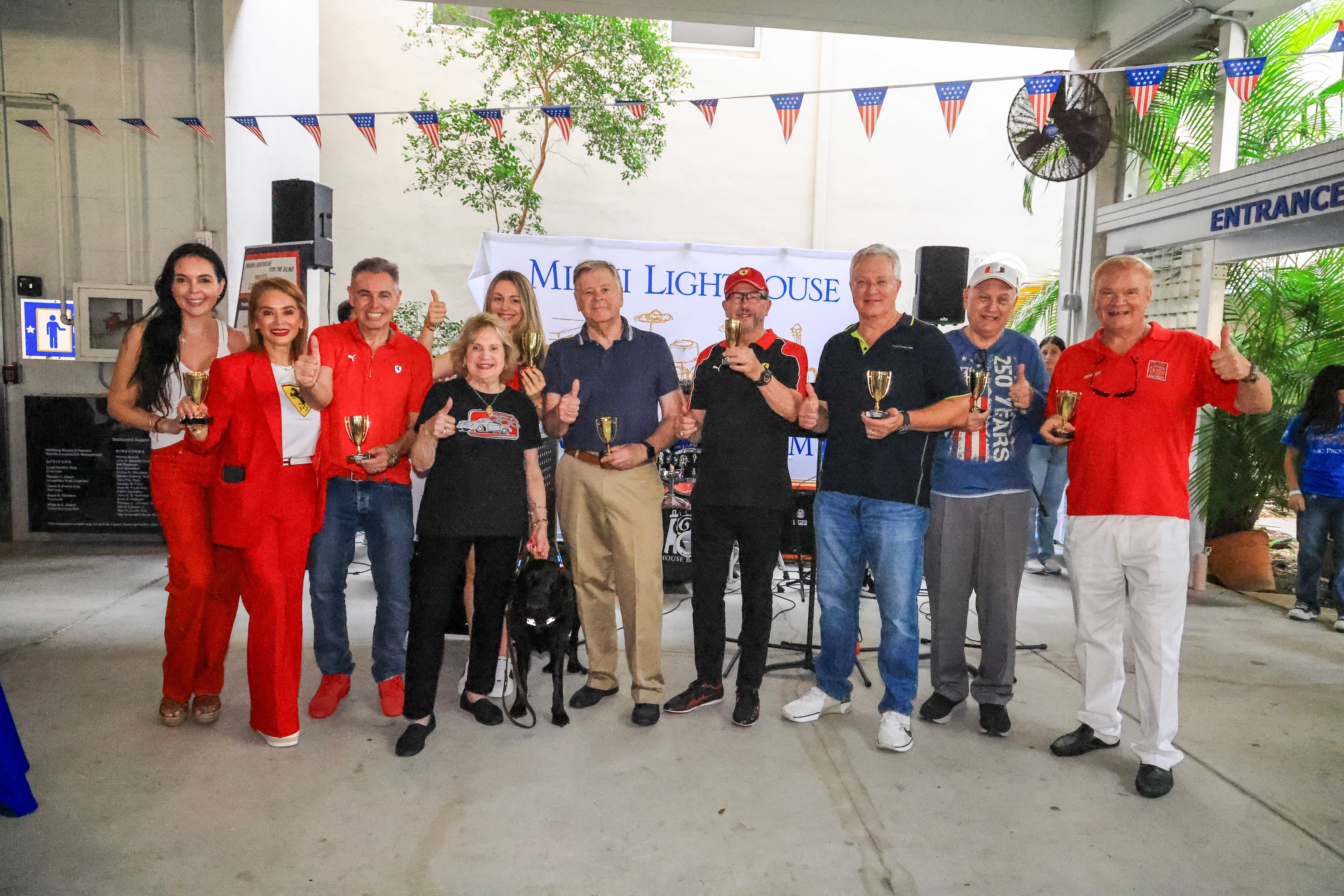 Sponsors and Miami Lighthouse leaders holding small gold trophies and giving a thumbs-up under American flag bunting, a black guide dog at the front