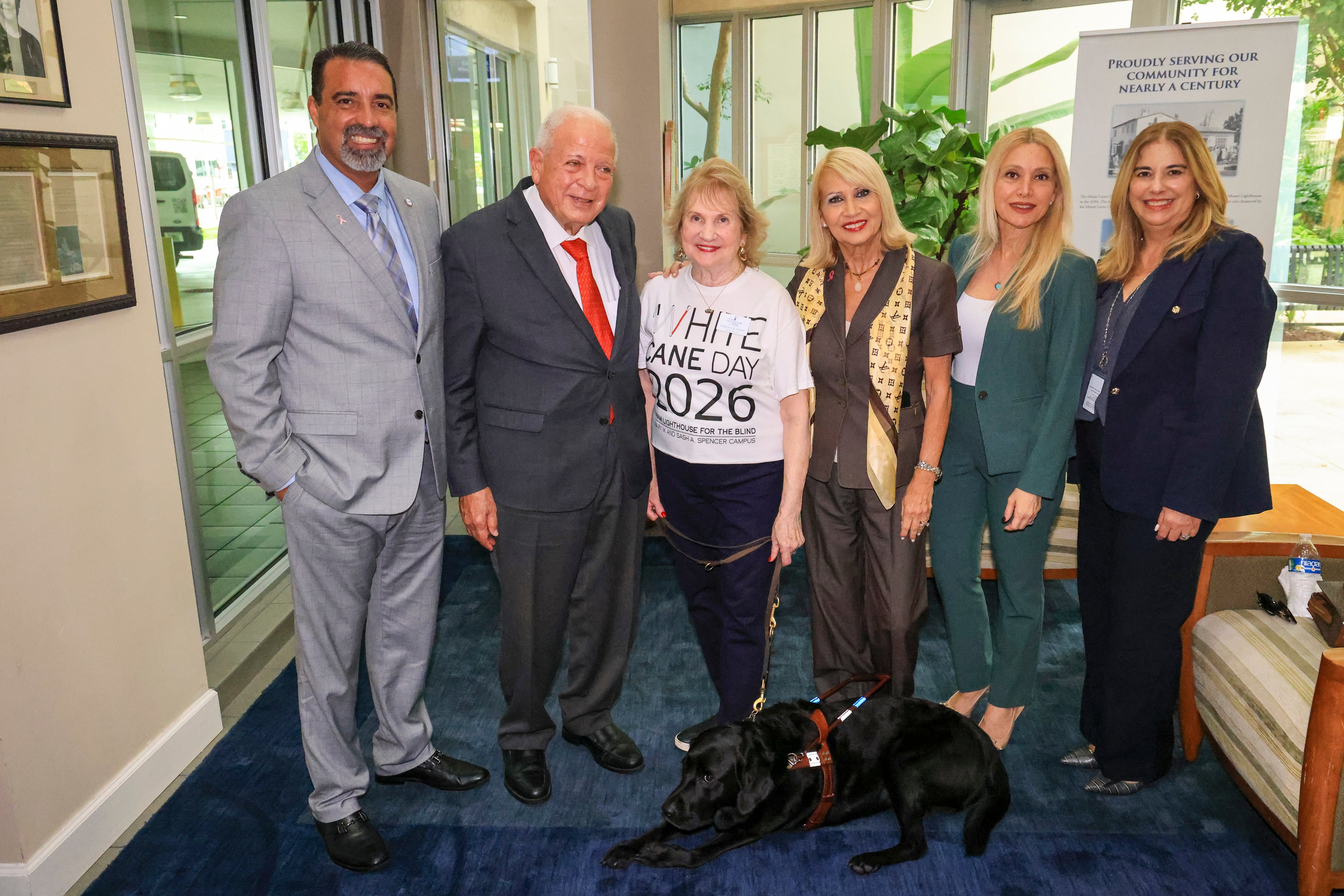 Seis invitados de pie en un interior, con un perro guía negro con arnés echado a sus pies