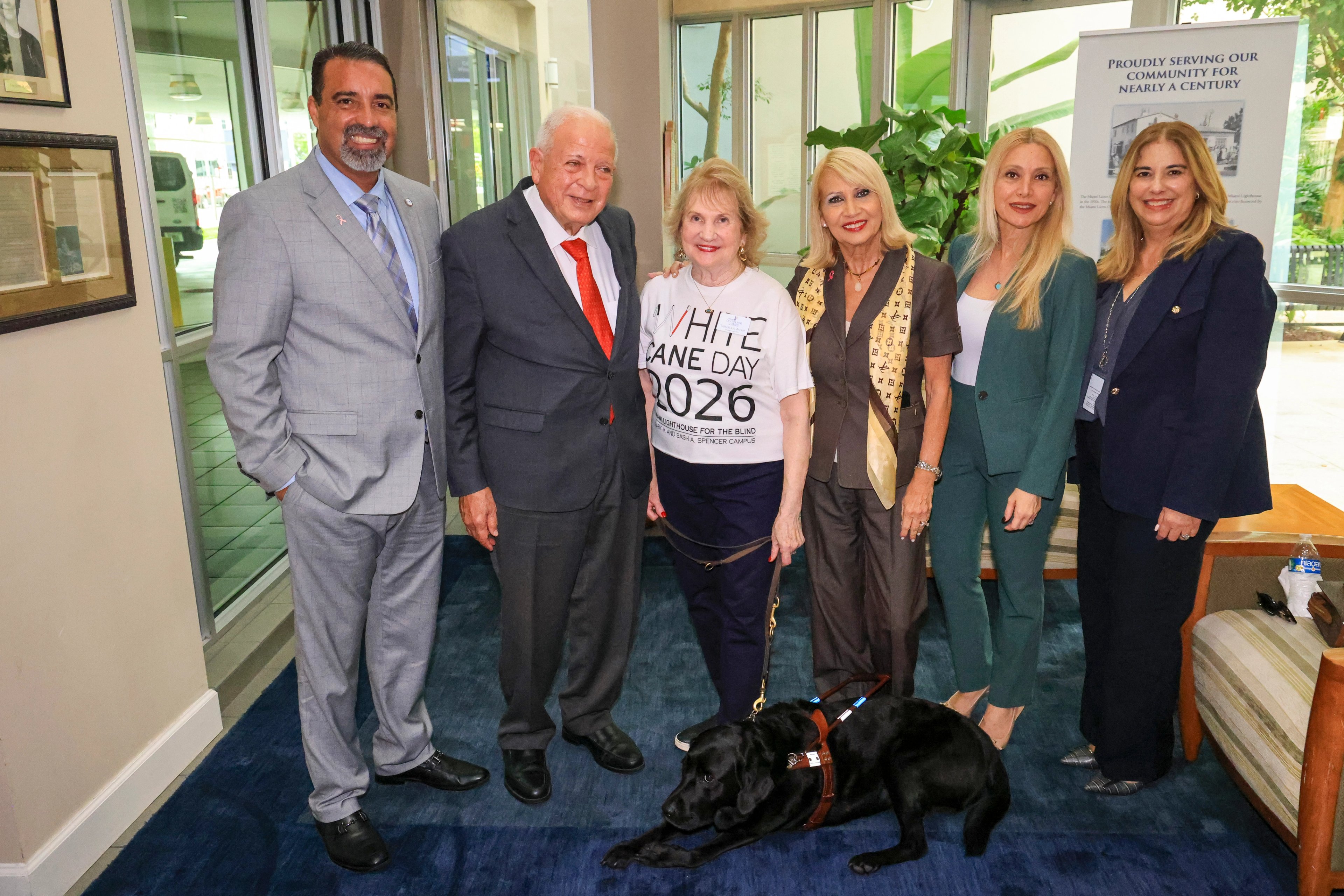 Six guests standing together indoors, a black guide dog in harness lying at their feet