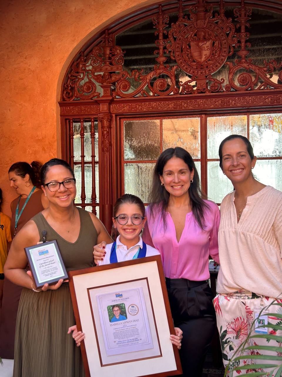 Una niña sostiene su reconocimiento enmarcado y sonríe junto a tres mujeres frente a una puerta en arco ornamentada
