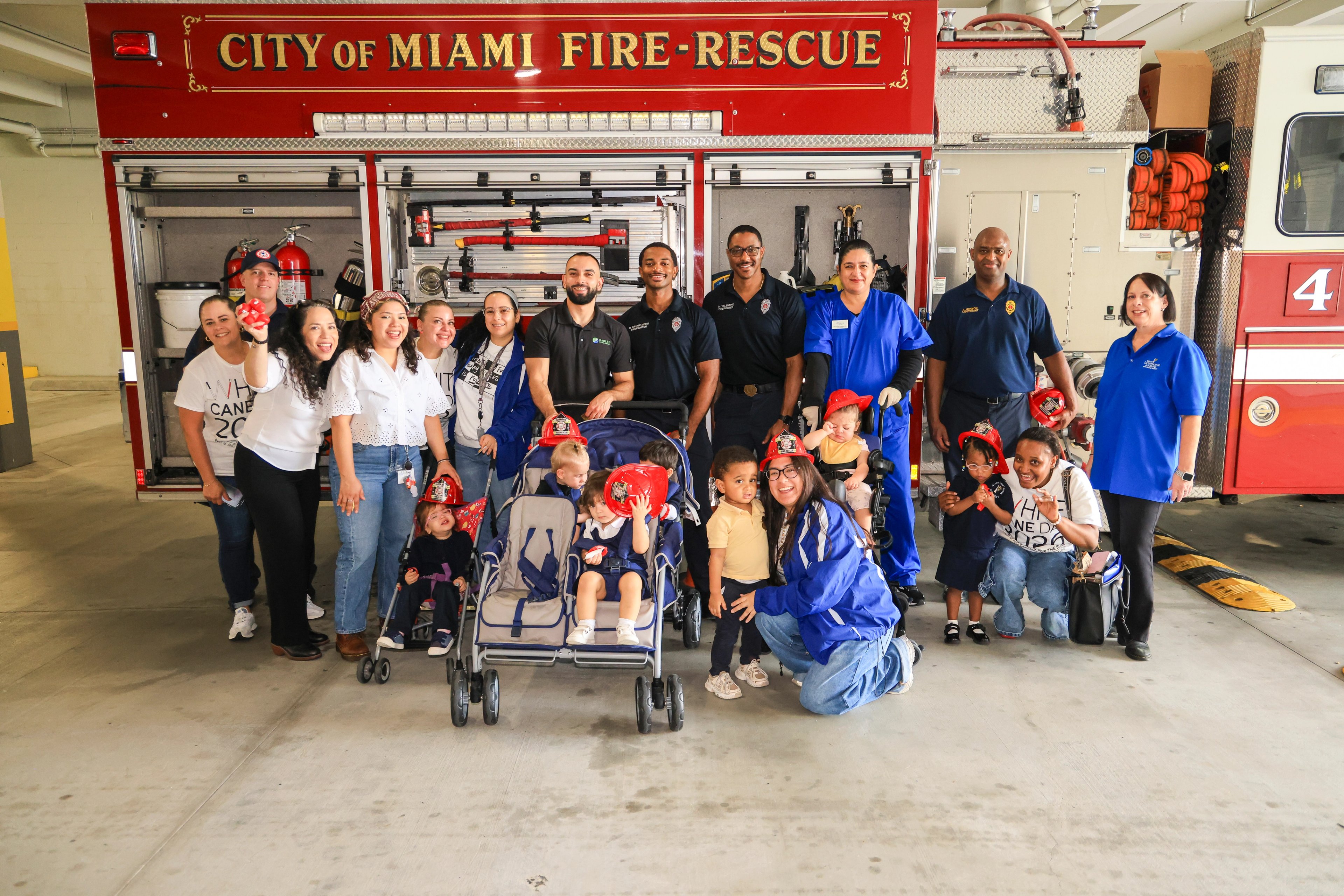Firefighters and other adults with toddlers in strollers and red toy fire helmets in front of a City of Miami Fire-Rescue truck