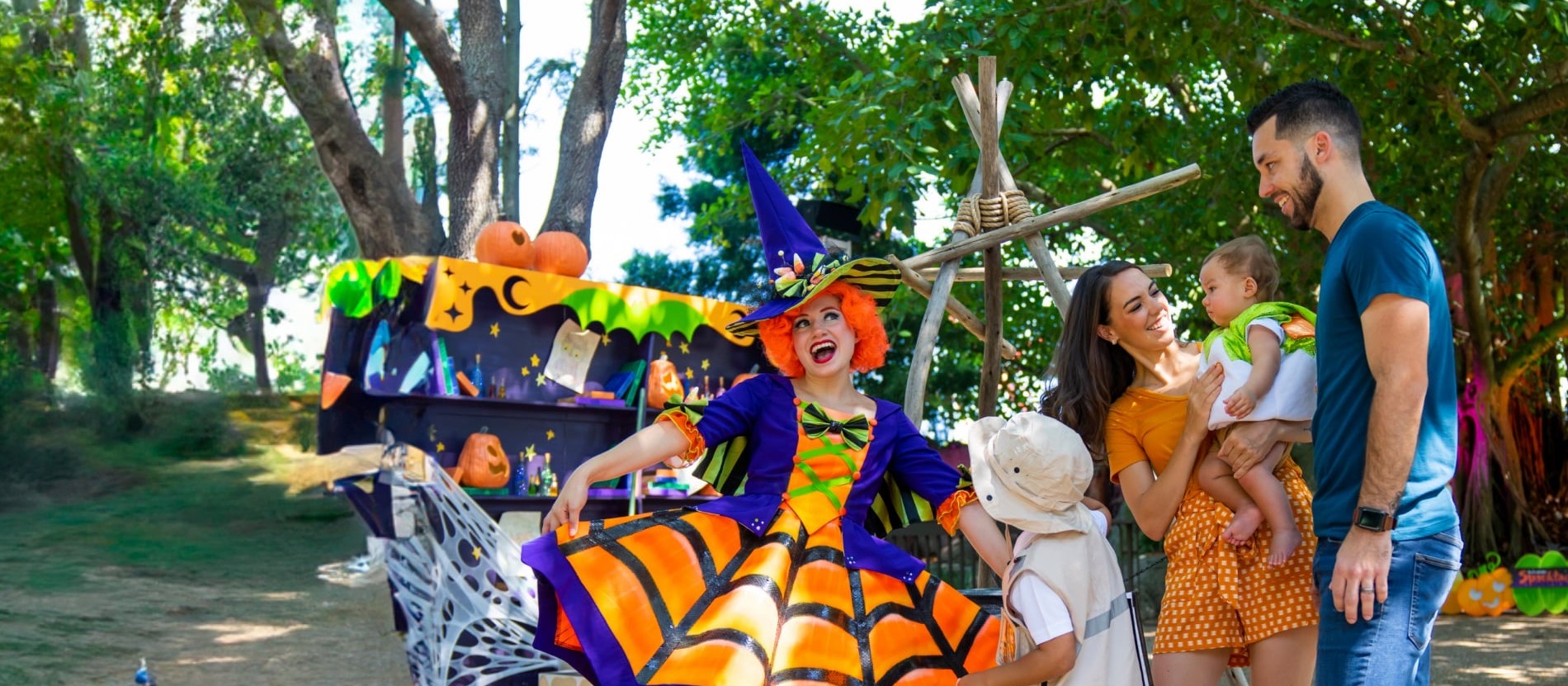 A witch in an orange-and-purple costume chatting with a family and a little boy in a safari hat under the trees at Busch Gardens