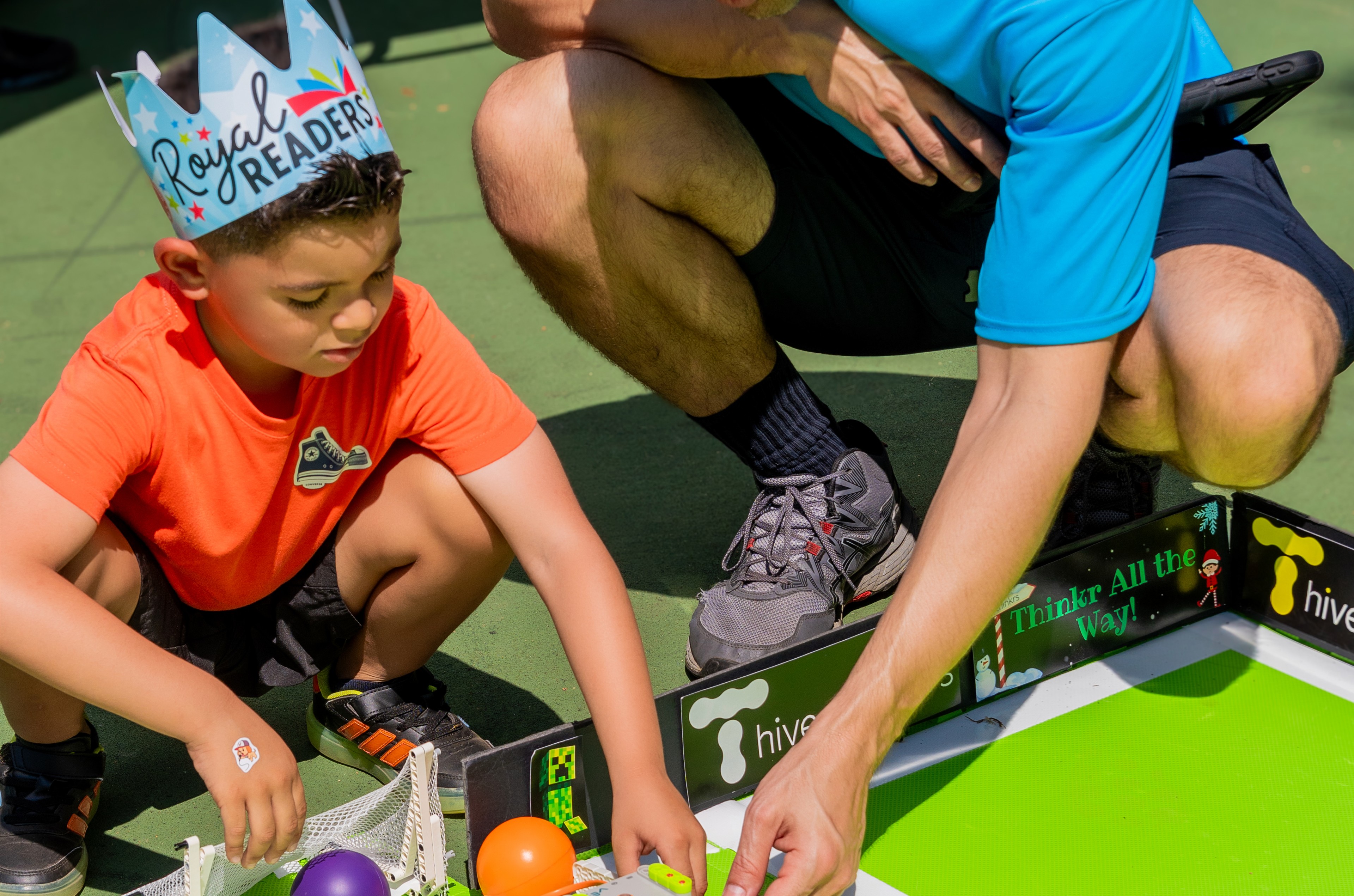 Un niño con una corona de papel arma un robot junto a un instructor de Hive of Thinkrs en un evento al aire libre