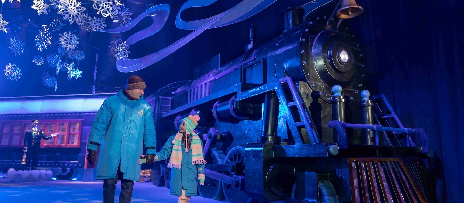 A parent and child in blue parkas walking past an ice-carved Polar Express locomotive