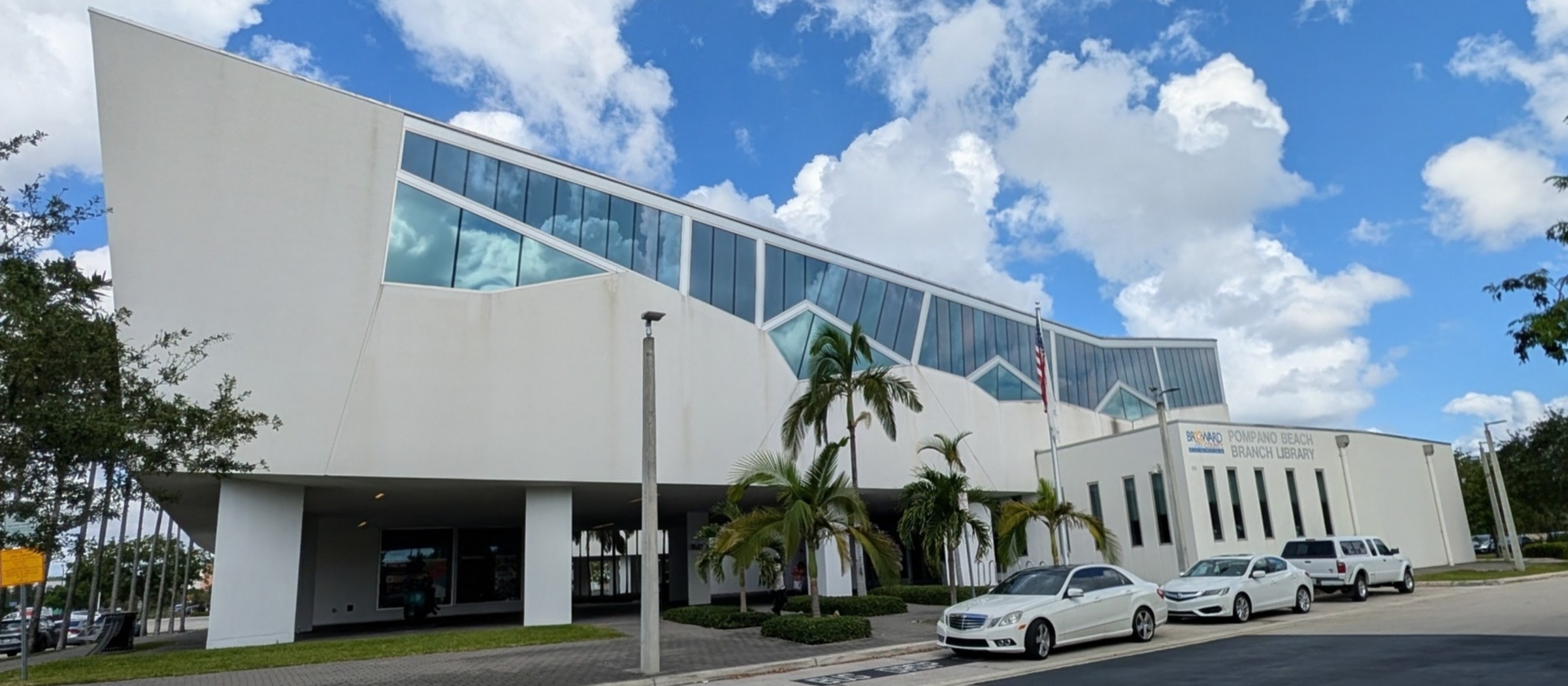 The Pompano Beach Library and Cultural Center, a white building with a sweeping curved wall and a band of tall windows, under a blue sky with clouds