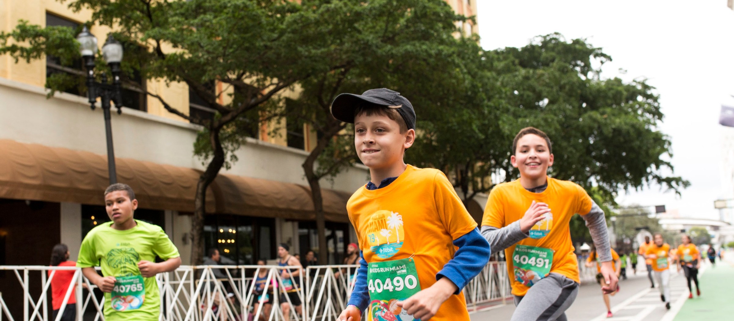Kids in race bibs and orange and green shirts run down a downtown Miami street