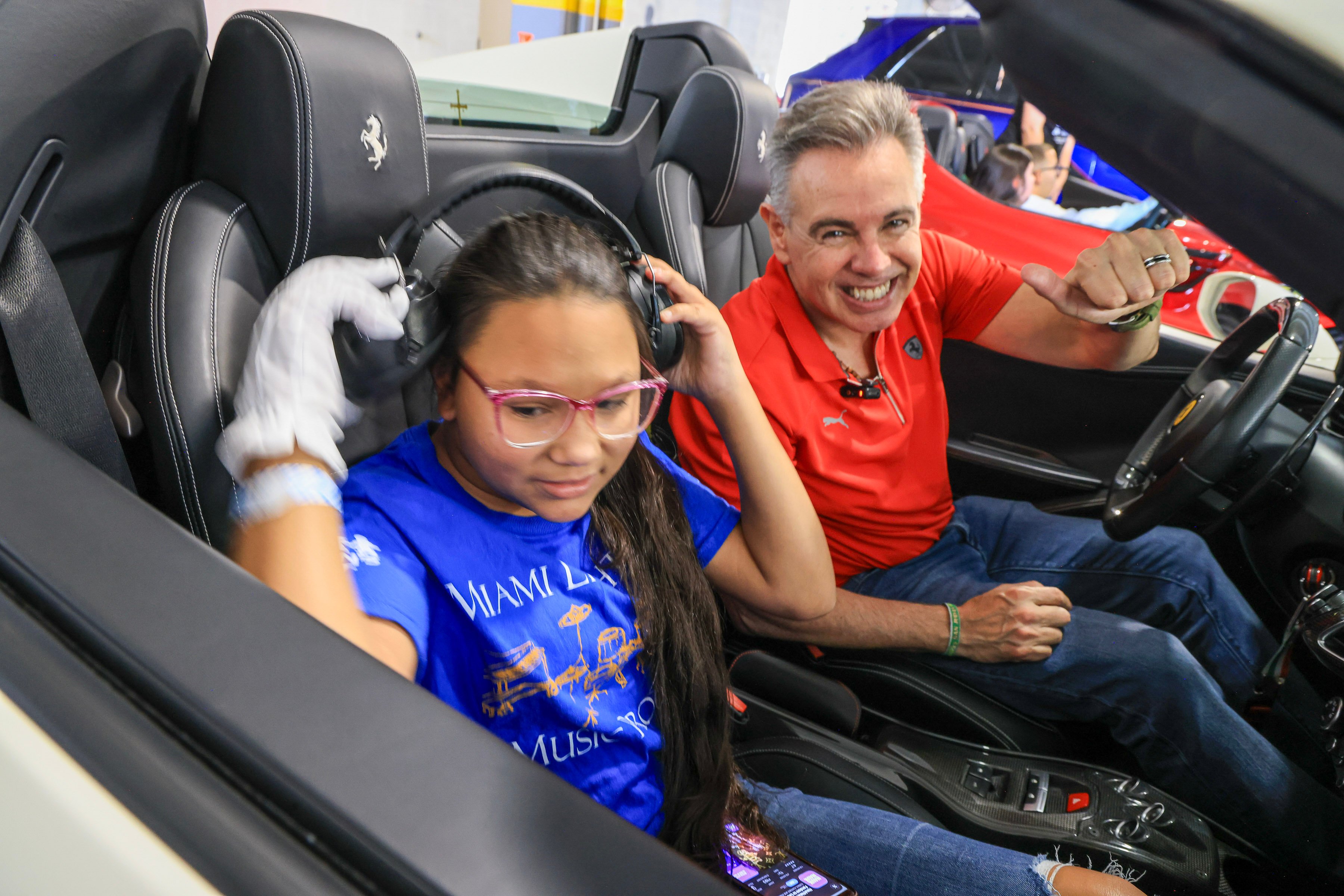 A student in headphones and white gloves in the passenger seat of a Ferrari convertible, the driver beside her smiling with a thumbs-up