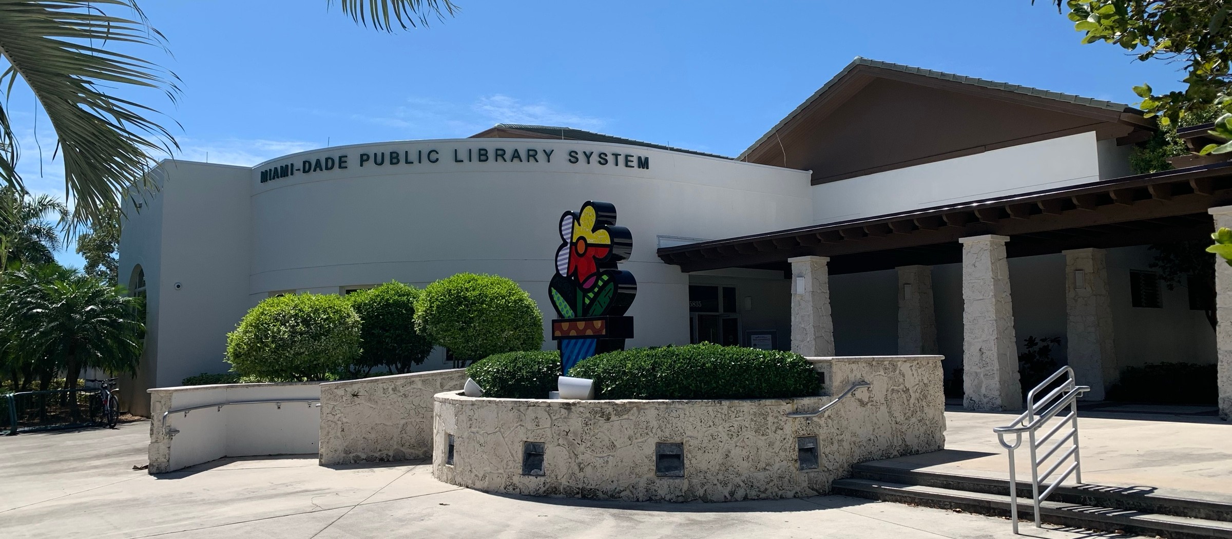 The white curved facade of the Pinecrest Branch Library with a colorful sculpture out front