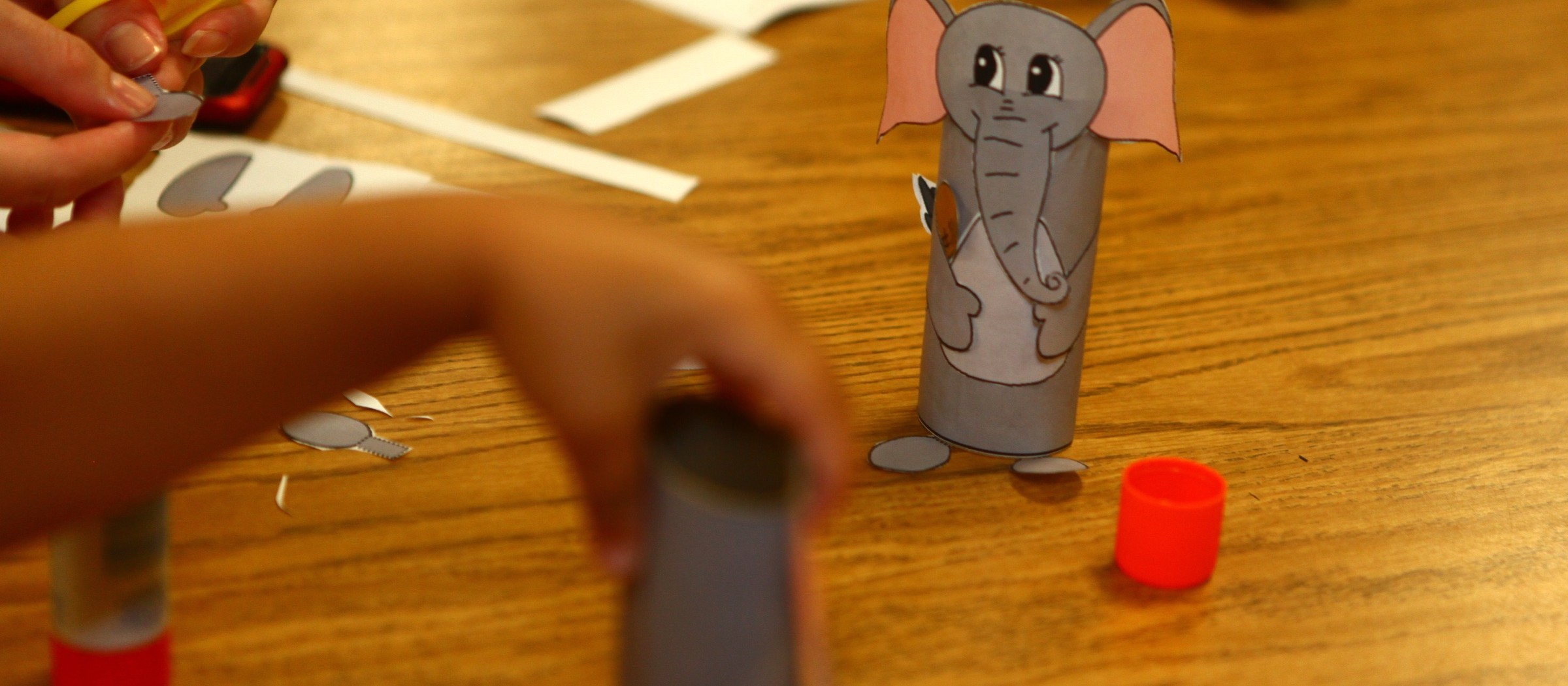 Un elefante hecho con un tubo de cartón sobre una mesa de biblioteca, con manos de niños trabajando al lado