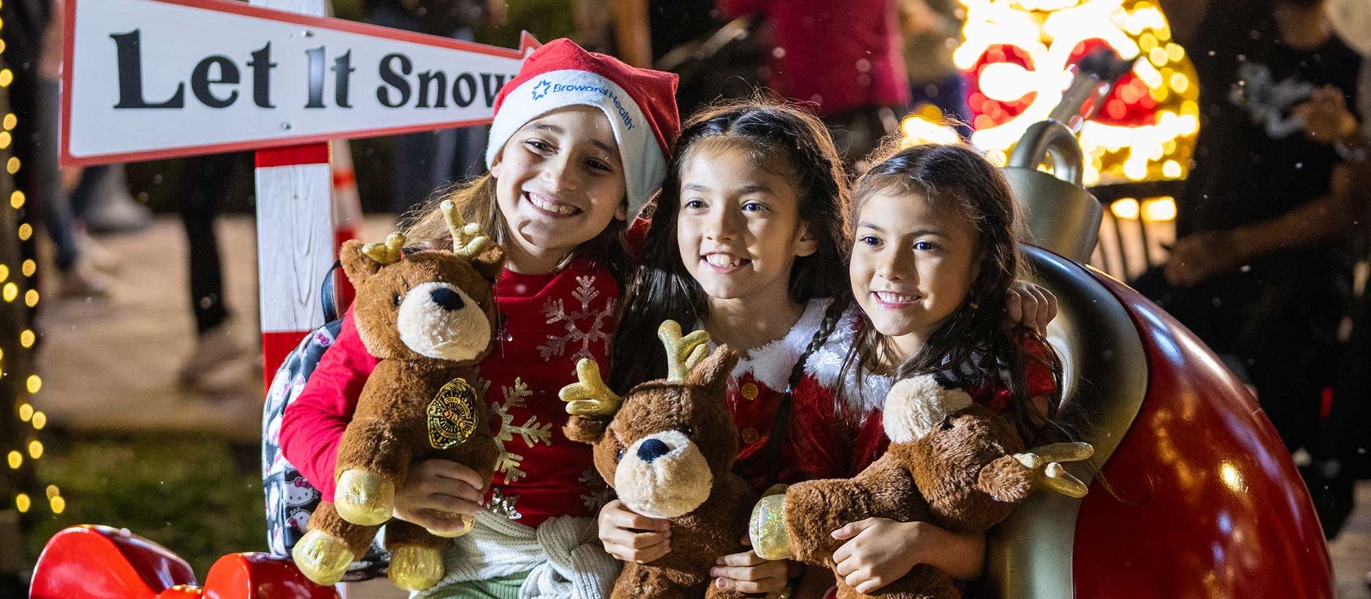 Three smiling girls in holiday outfits holding reindeer plush toys beside a giant ornament and a 'Let it Snow' sign