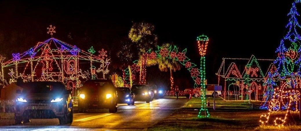 Autos con los faros encendidos pasan junto a figuras de luces de una casa de jengibre, palmeras y un farol de noche
